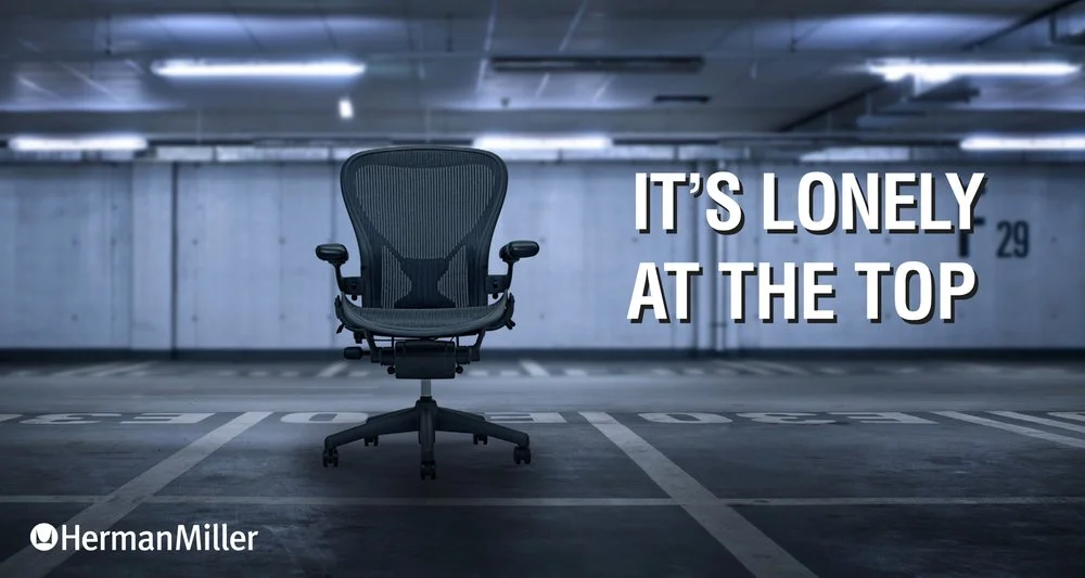 An empty office chair in a dimly lit parking garage with the text 'IT'S LONELY AT THE TOP' and Herman Miller branding.