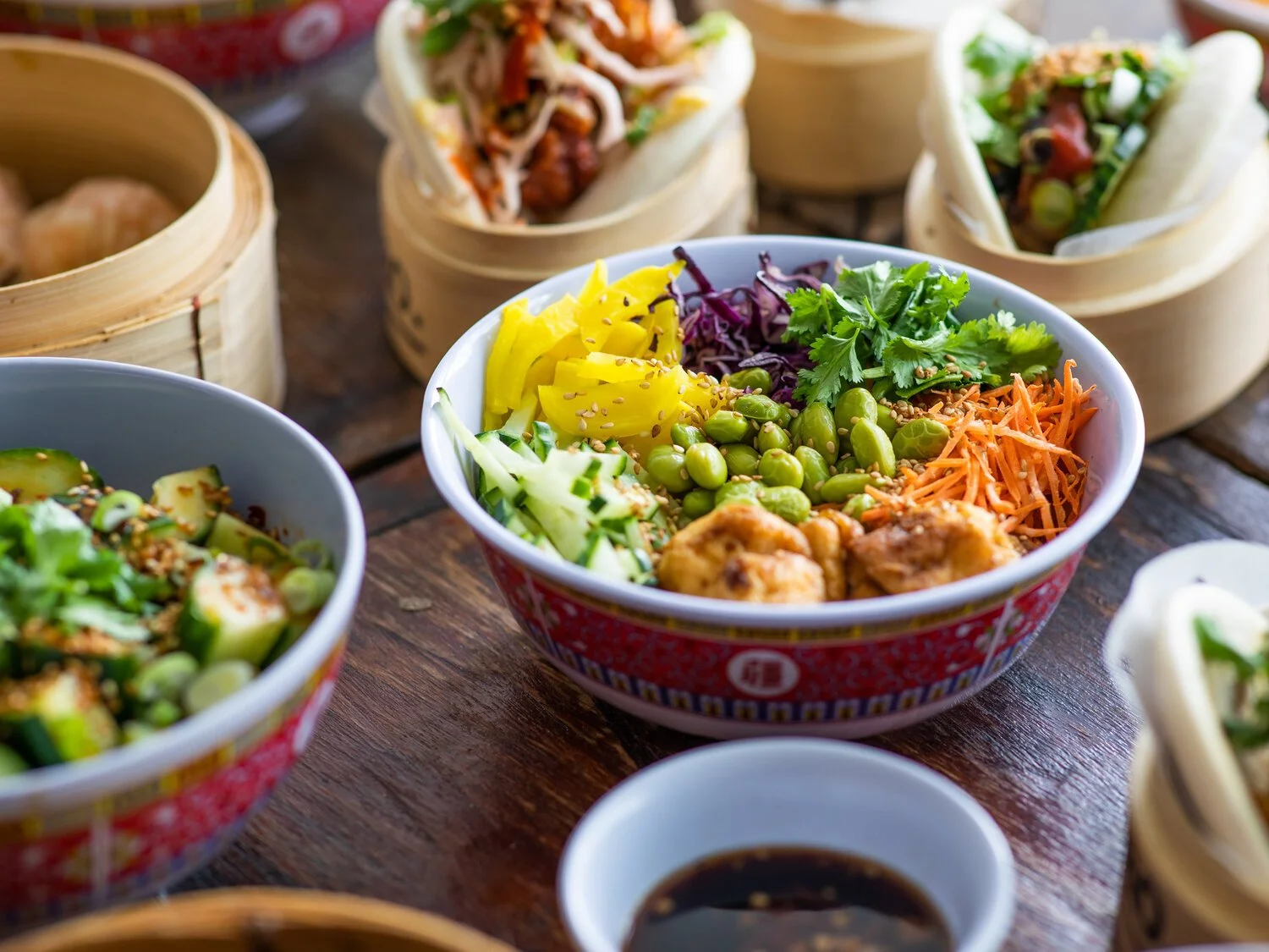 A bowl of colorful Asian salad with shredded carrots, cucumber, purple cabbage, edamame, yellow bell peppers, cilantro, and chicken pieces, surrounded by various dim sum dishes and soy sauce on a wooden table.