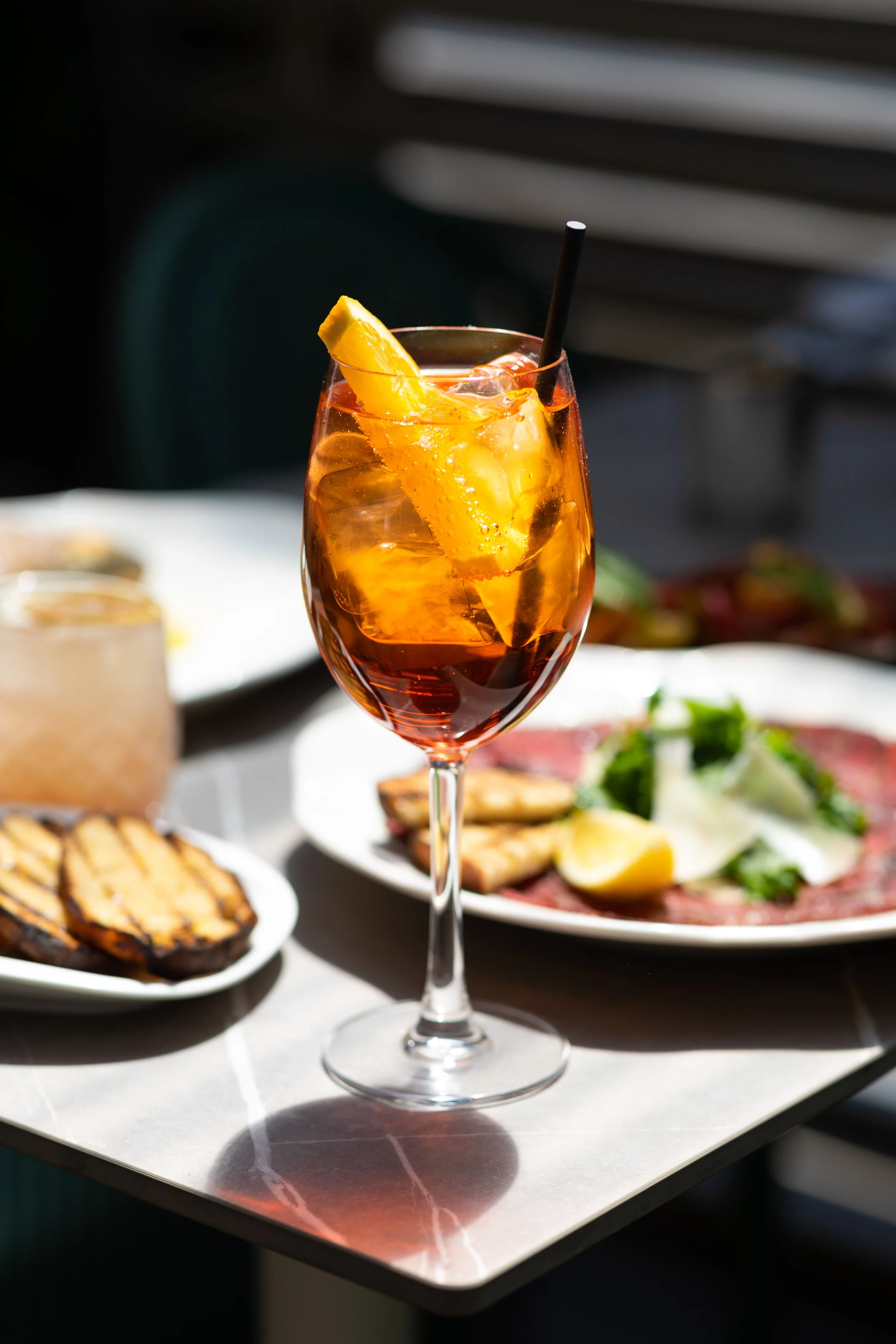 A glass of iced Aperol spritz with orange slice on a table with food in the background