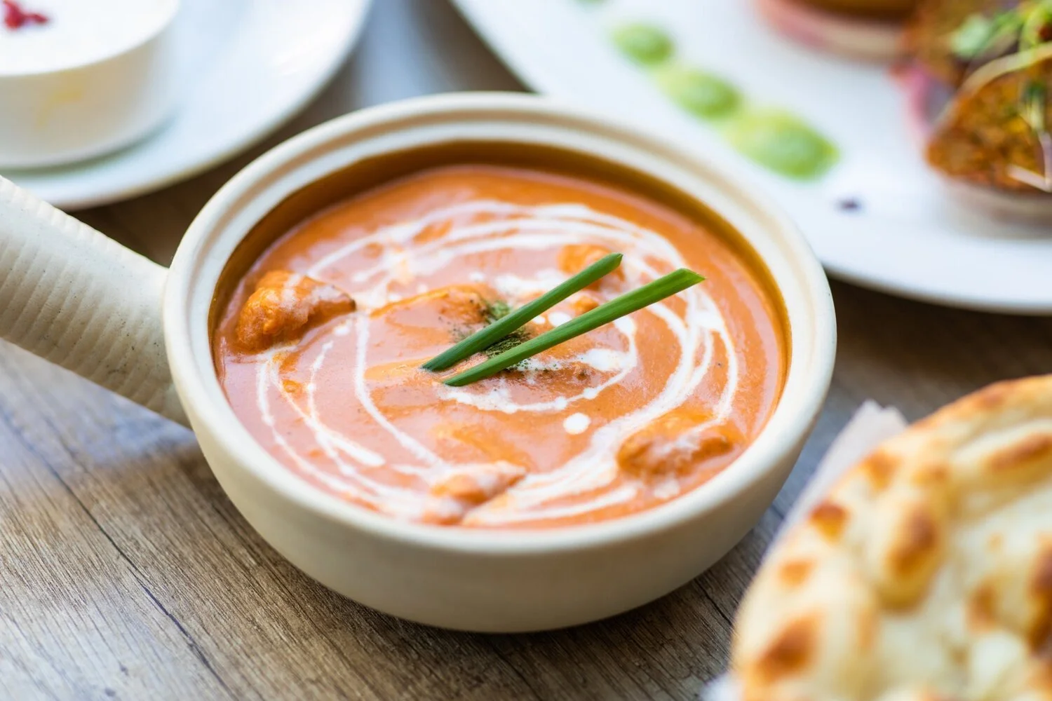 Close-up of a bowl of creamy tomato soup garnished with two green chives and a swirl of cream, served with bread on a wooden table.