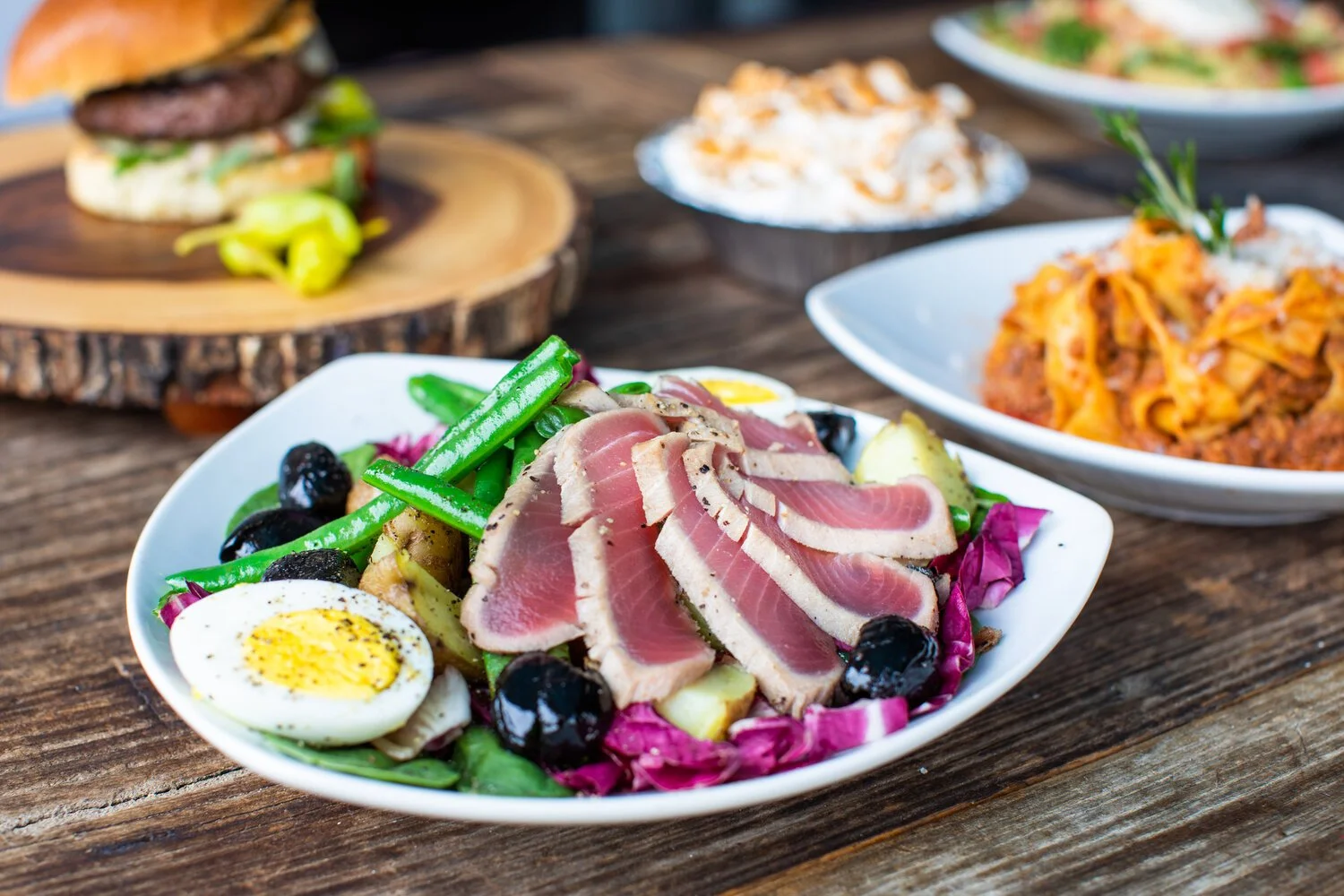 A plate of salad with sliced tuna, boiled egg, green beans, black olives, and purple cabbage on a rustic wooden table with other dishes in the background.