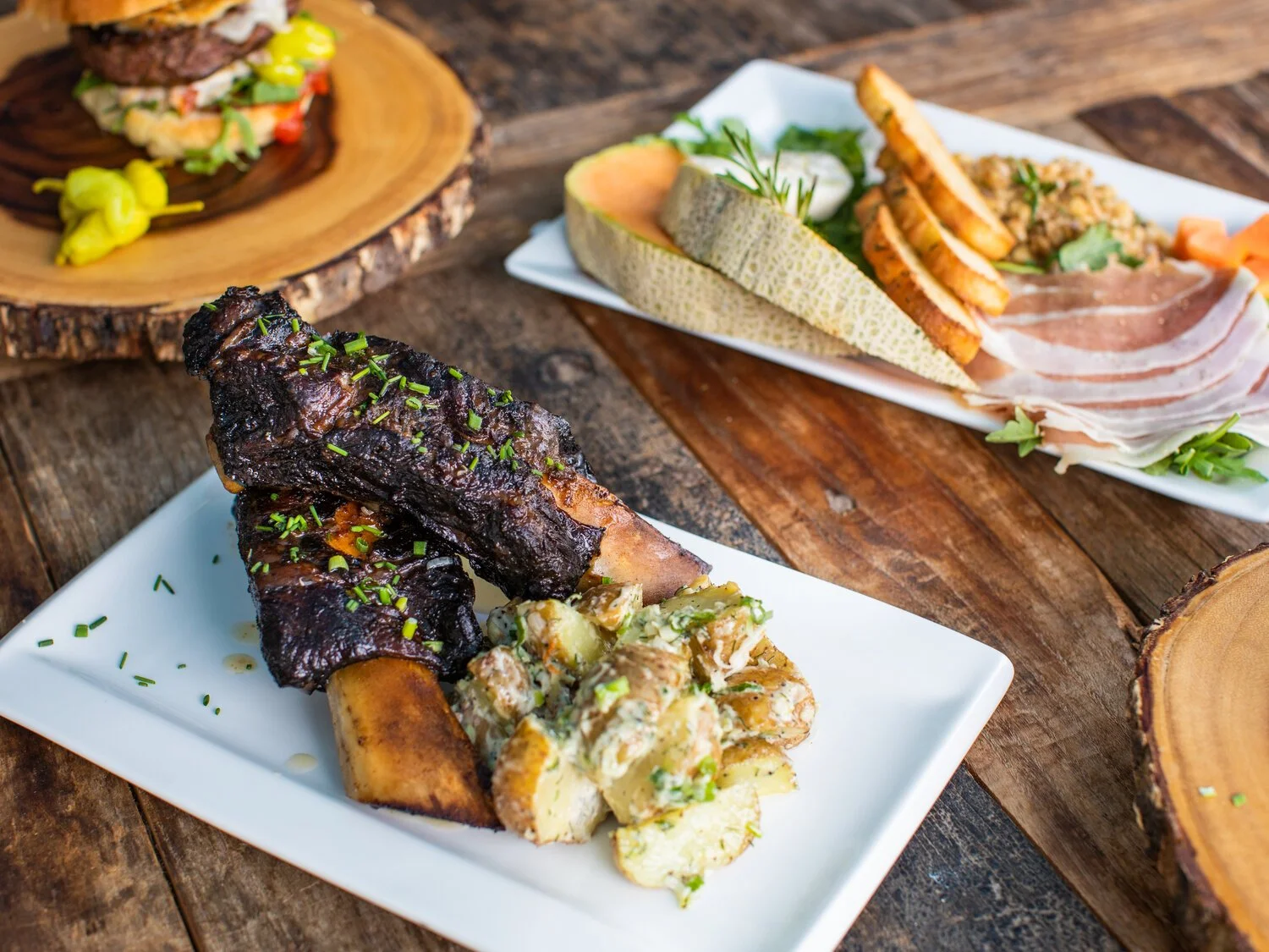 Barbecue ribs with potatoes and vegetables on a white plate, charcuterie with cheese, vegetables, and sliced meat on a white tray, and a burger with toppings on a wooden serving board.