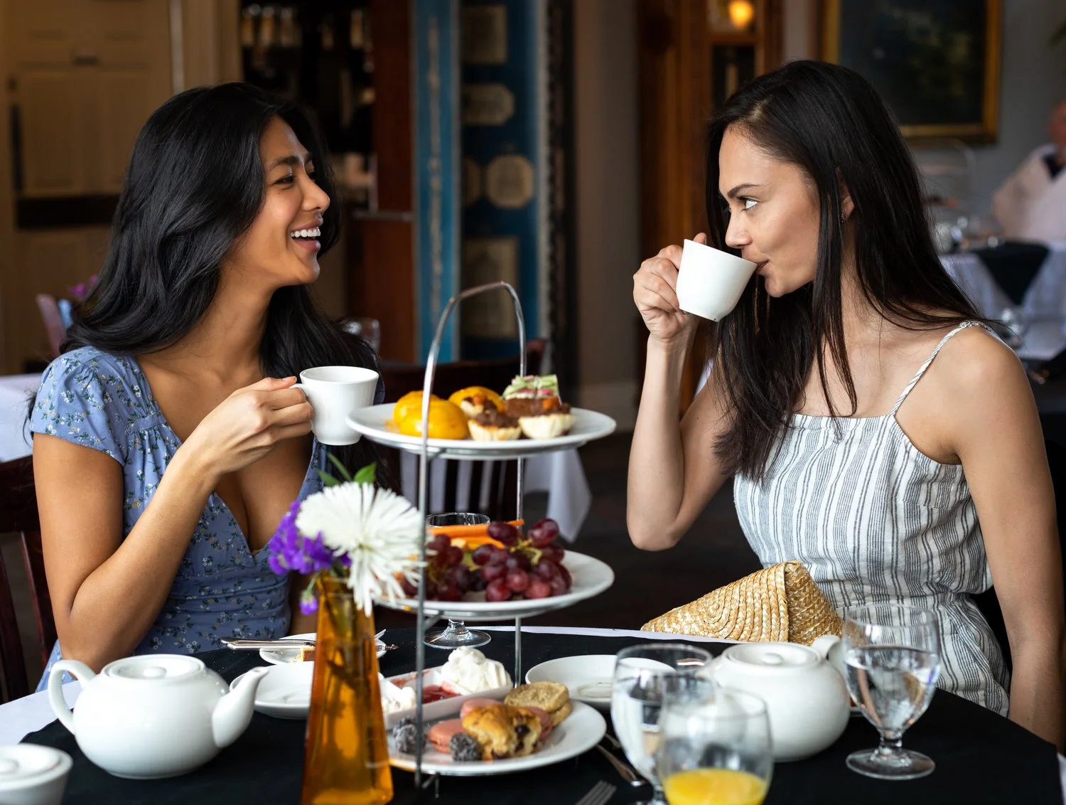 Two women enjoying tea and desserts at a restaurant table, smiling and engaging with each other, with assorted pastries and a flower arrangement on the table.