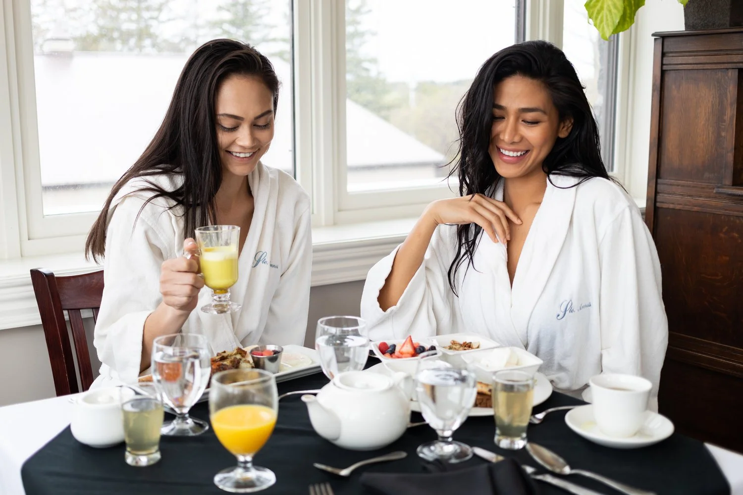 Two women in white bathrobes enjoying breakfast at a dining table with tea, juice, fruit, and baked goods, sitting by large windows with a view outside.