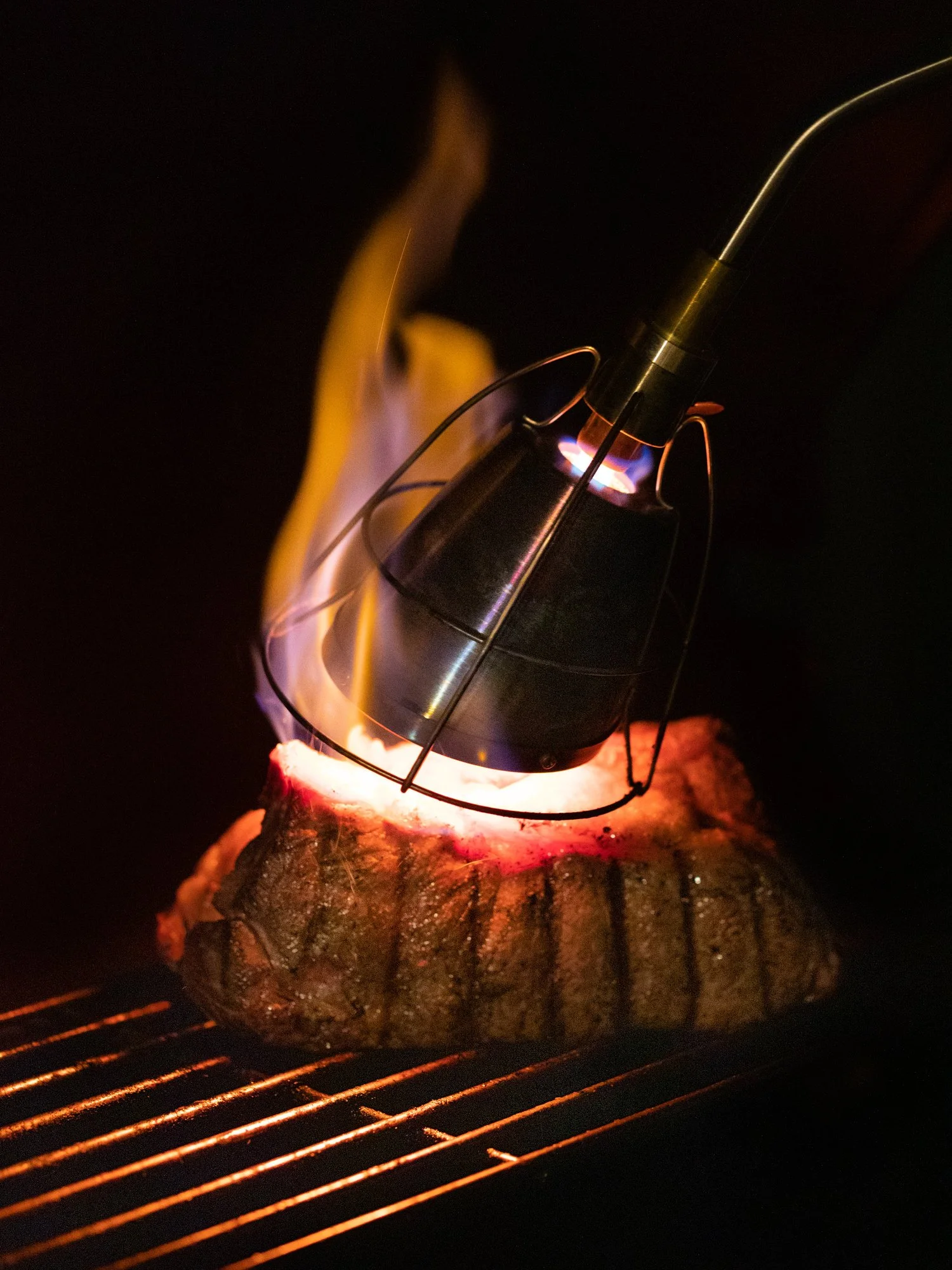 A meat probe thermometer is inserted into a steak on a grill, with flames and smoke visible around the meat.