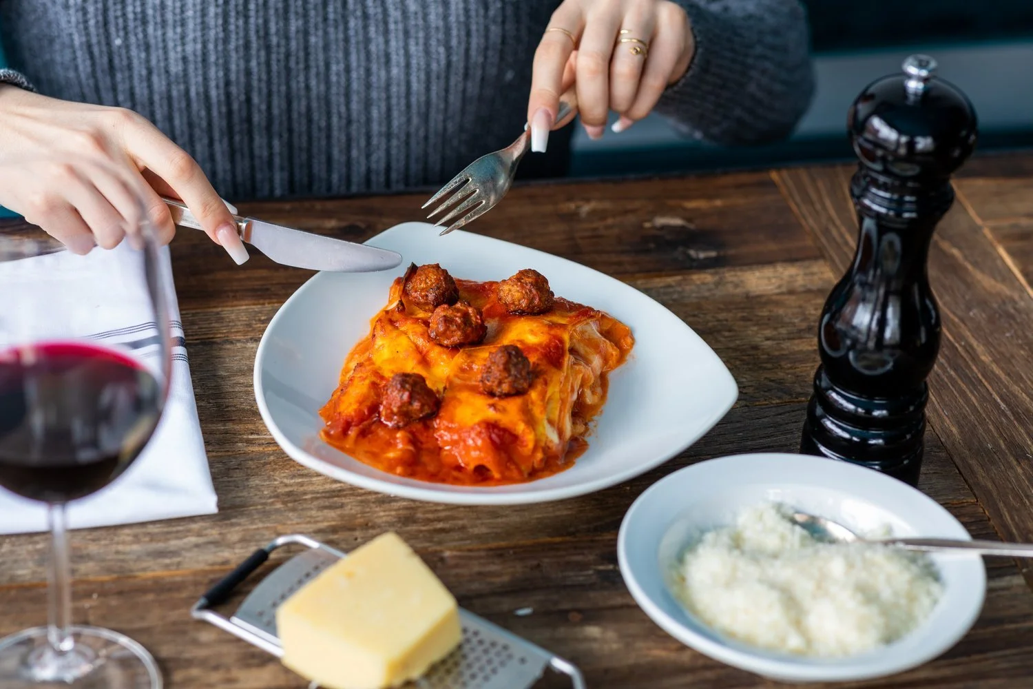 Person about to eat a slice of lasagna with meatballs on top, served on a white plate, with a glass of red wine, a bowl of grated cheese, and a salt grinder on a wooden table.