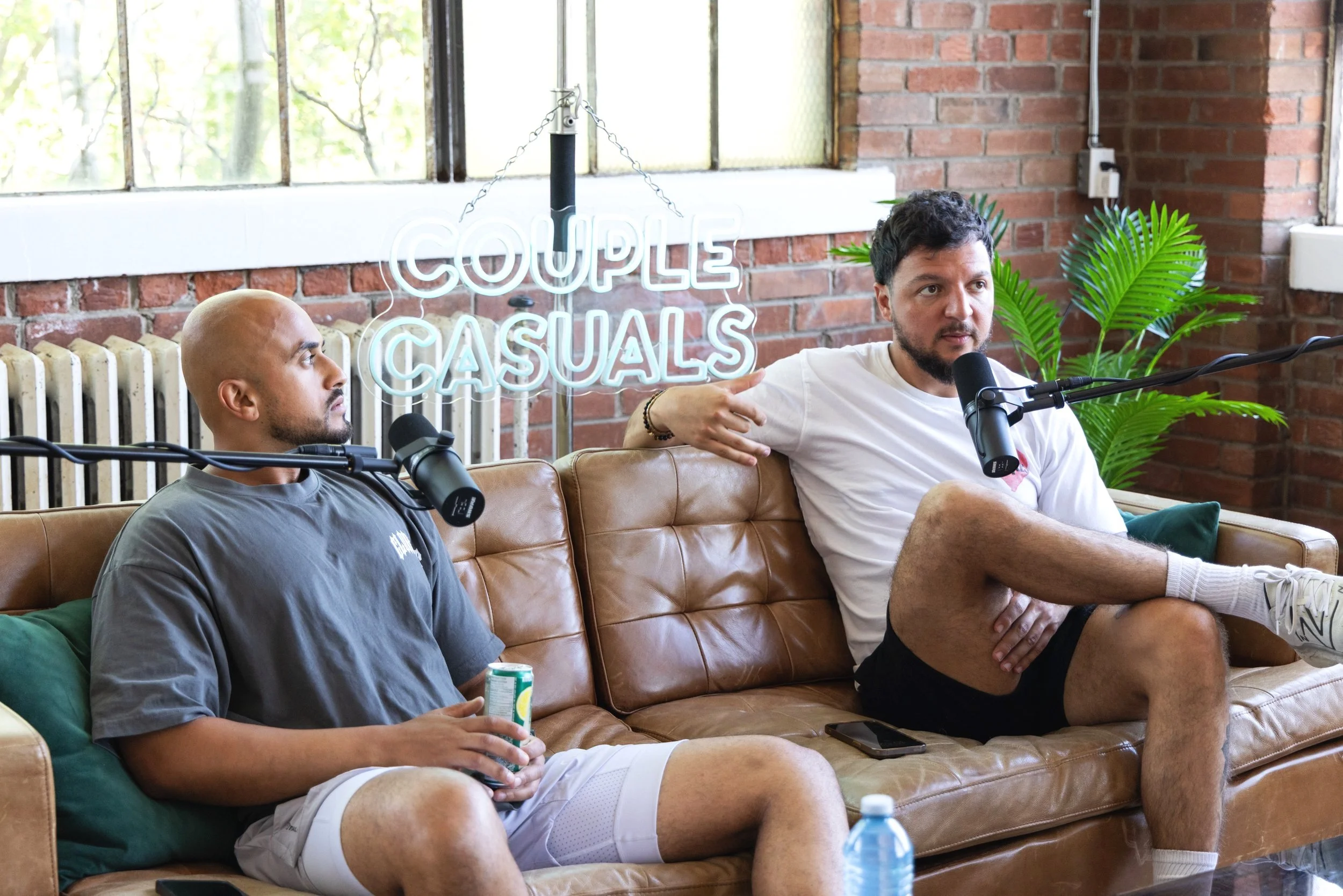 Two men sitting on a brown leather couch in a room with brick walls, engaged in a conversation with microphones in front of them. One man is bald and holding a can, the other has dark hair and is gesturing while speaking. Behind them is a neon sign that reads 'COUPLES CASUALS' and a green plant.