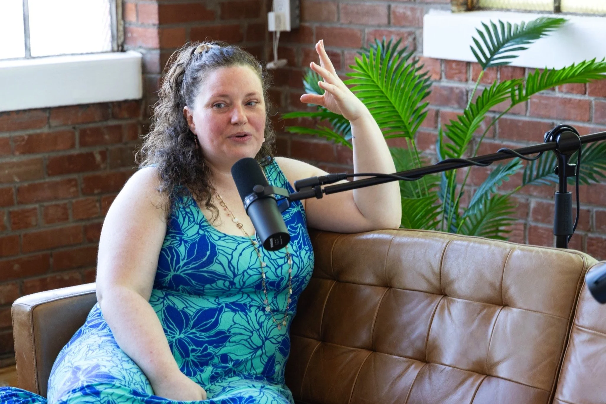 A woman with curly brown hair wearing a blue floral dress and gold necklace speaking into a microphone, sitting on a tan leather couch in a room with exposed brick walls and a green plant in the background.