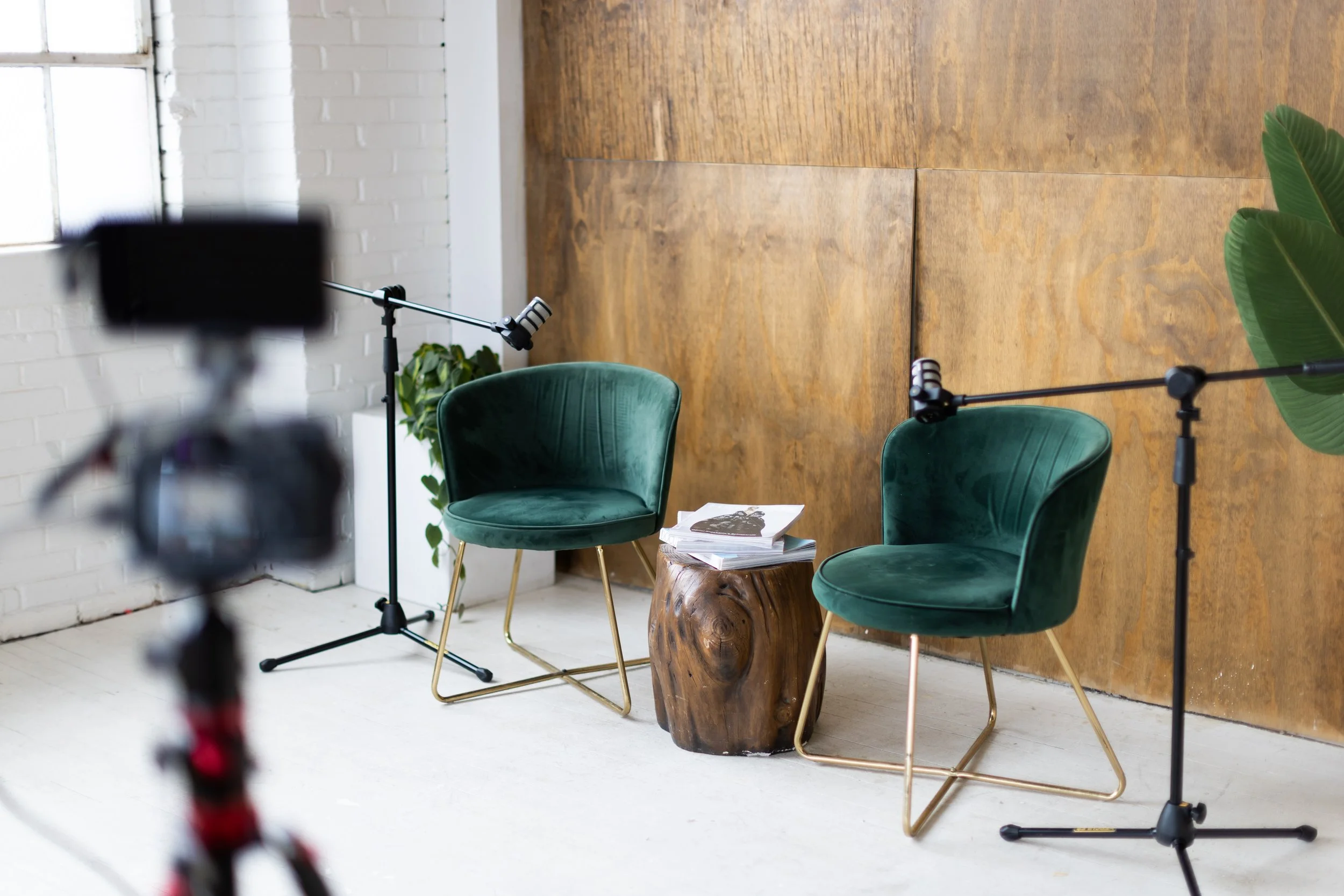Set up for an interview or podcast with two green velvet chairs with gold legs, a small wooden table with magazines, a potted plant in the corner, and two boom microphones on stands, inside a room with white brick and wood-paneled walls.