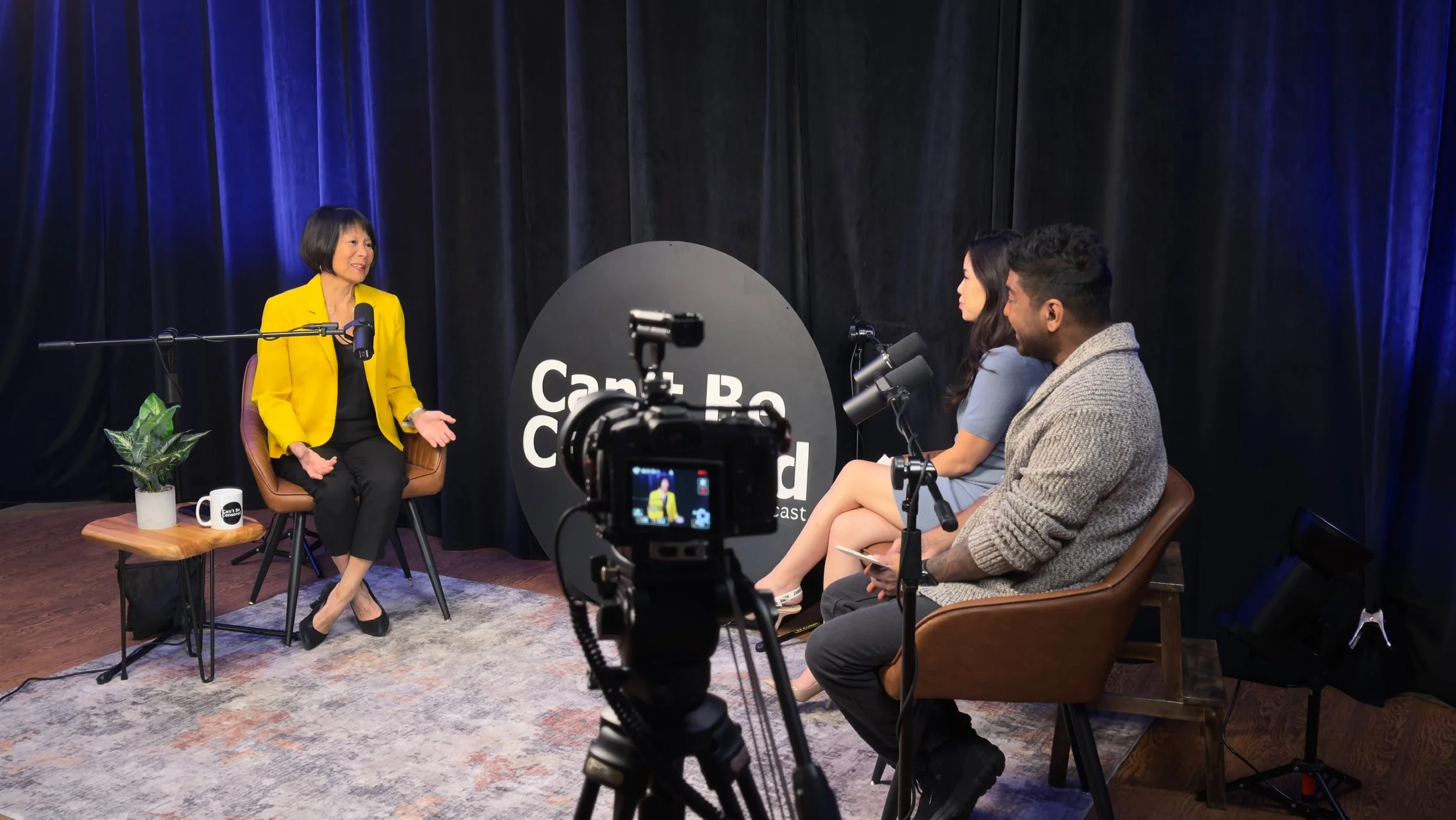 Two women and one man are seated on stage during an interview recording. The woman on the left, wearing a yellow blazer, is speaking, while the other two listen. A video camera is set up in front of them, capturing the scene. The background is black 