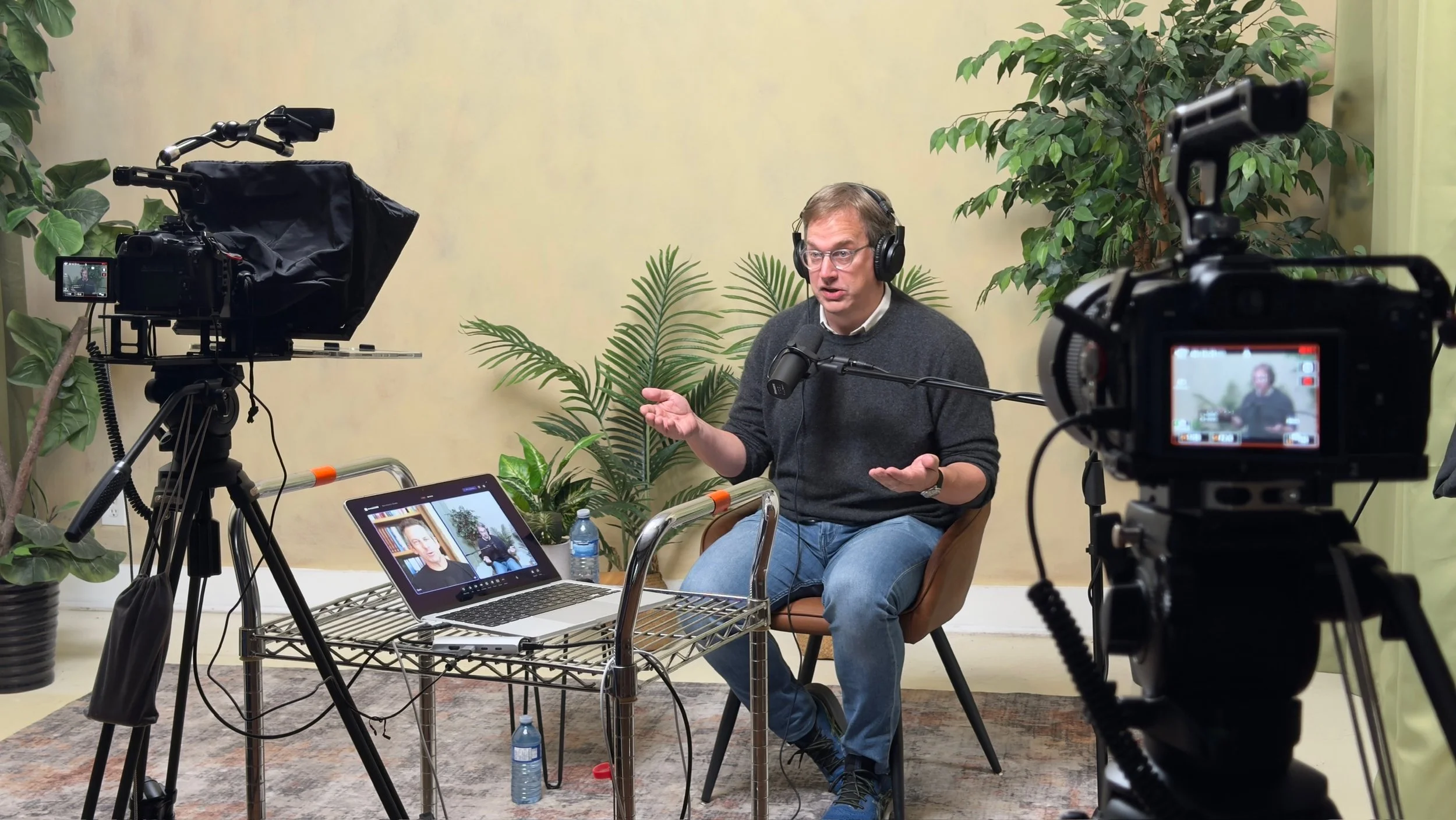 A man wearing glasses and headphones is sitting and speaking into a microphone during a video recording, with cameras and a laptop set up around him and green plants in the background.