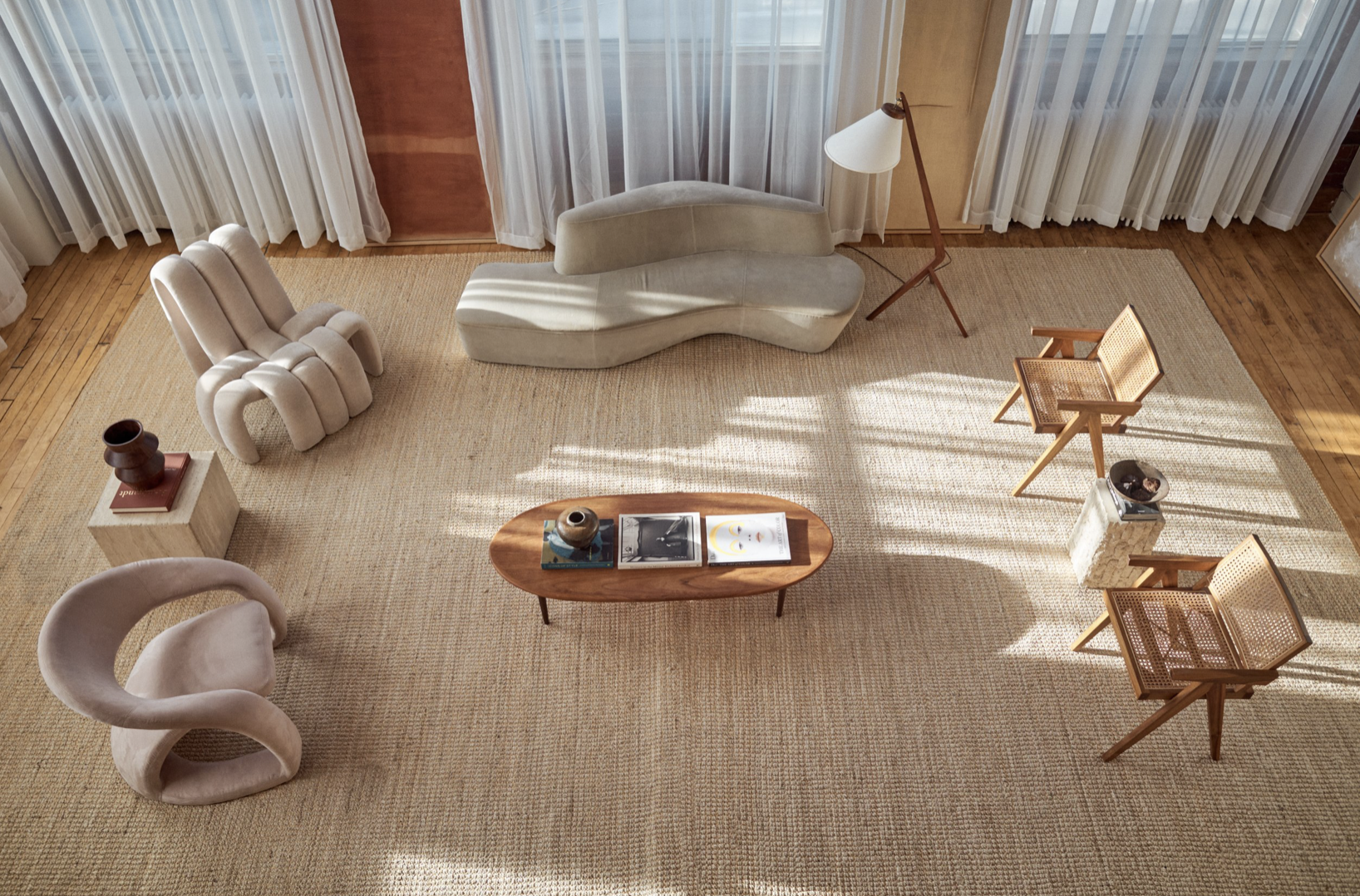 Living room with a beige armchair, a curved sofa, a wooden coffee table with books and vases, three wooden chairs, a standing lamp, and beige curtains with sunlight streaming inside.