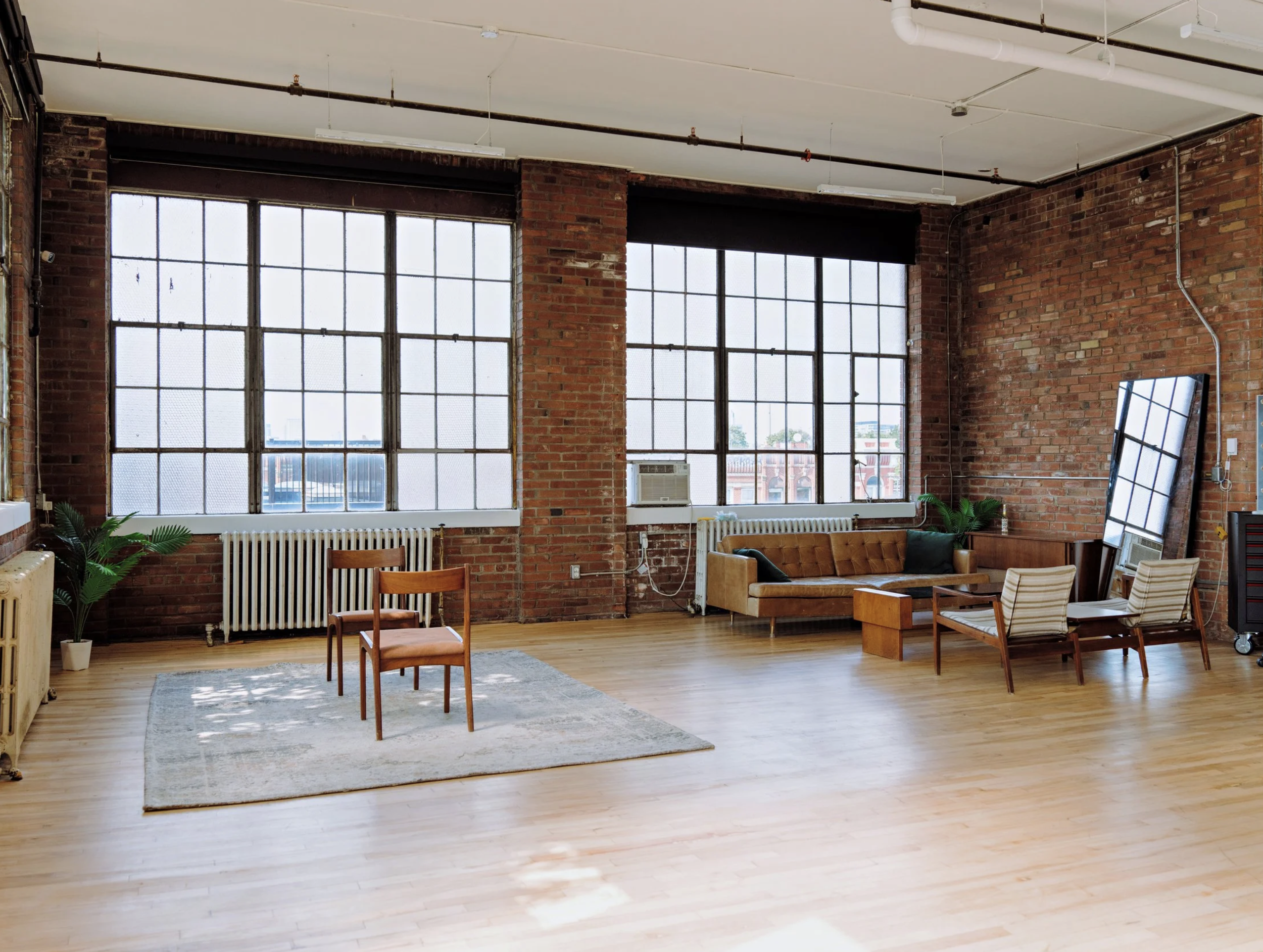 An industrial-style living room with large windows, brick walls, hardwood floors, a vintage sofa, chairs, a mirror, and indoor plants.