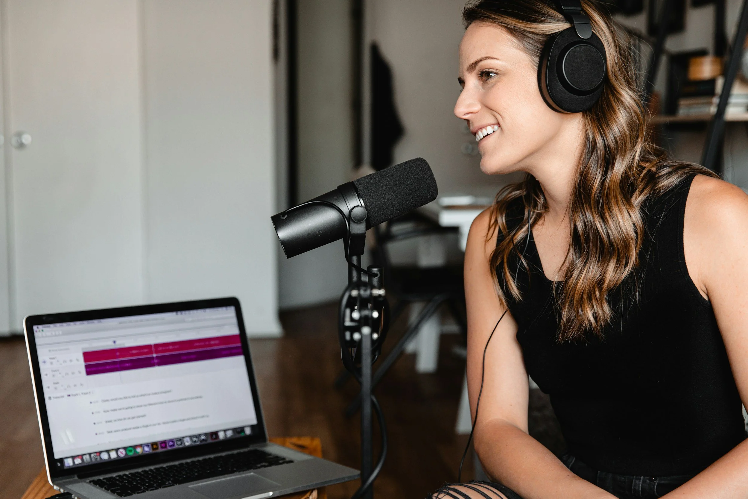 A woman with long wavy hair wearing a black tank top and headphones, speaking into a microphone, with a laptop open in front of her, in a home studio setting.