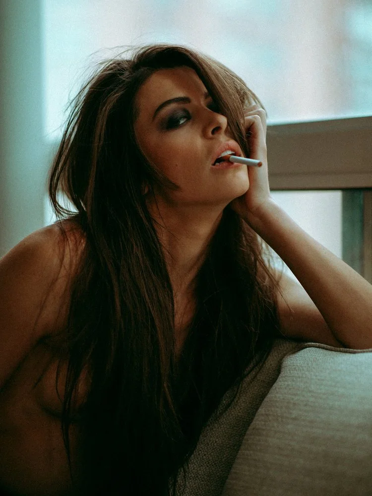 A woman with long dark hair and dark makeup, sitting on a couch, holding a cigarette in her mouth, resting her head on her hand, and looking thoughtfully out of a window.