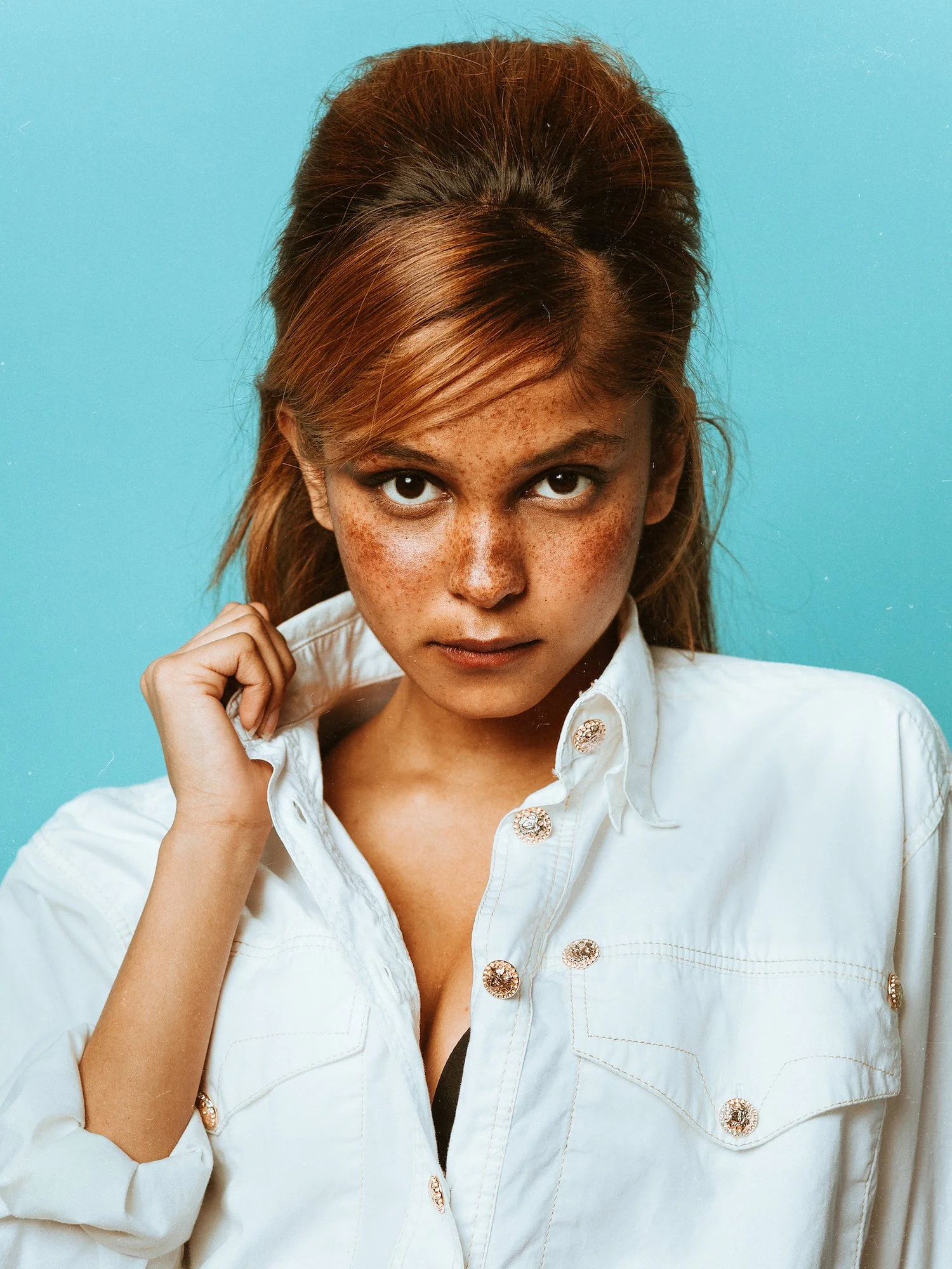 A woman with freckled skin and reddish-brown hair, wearing a partially unbuttoned white shirt and against a light blue background.