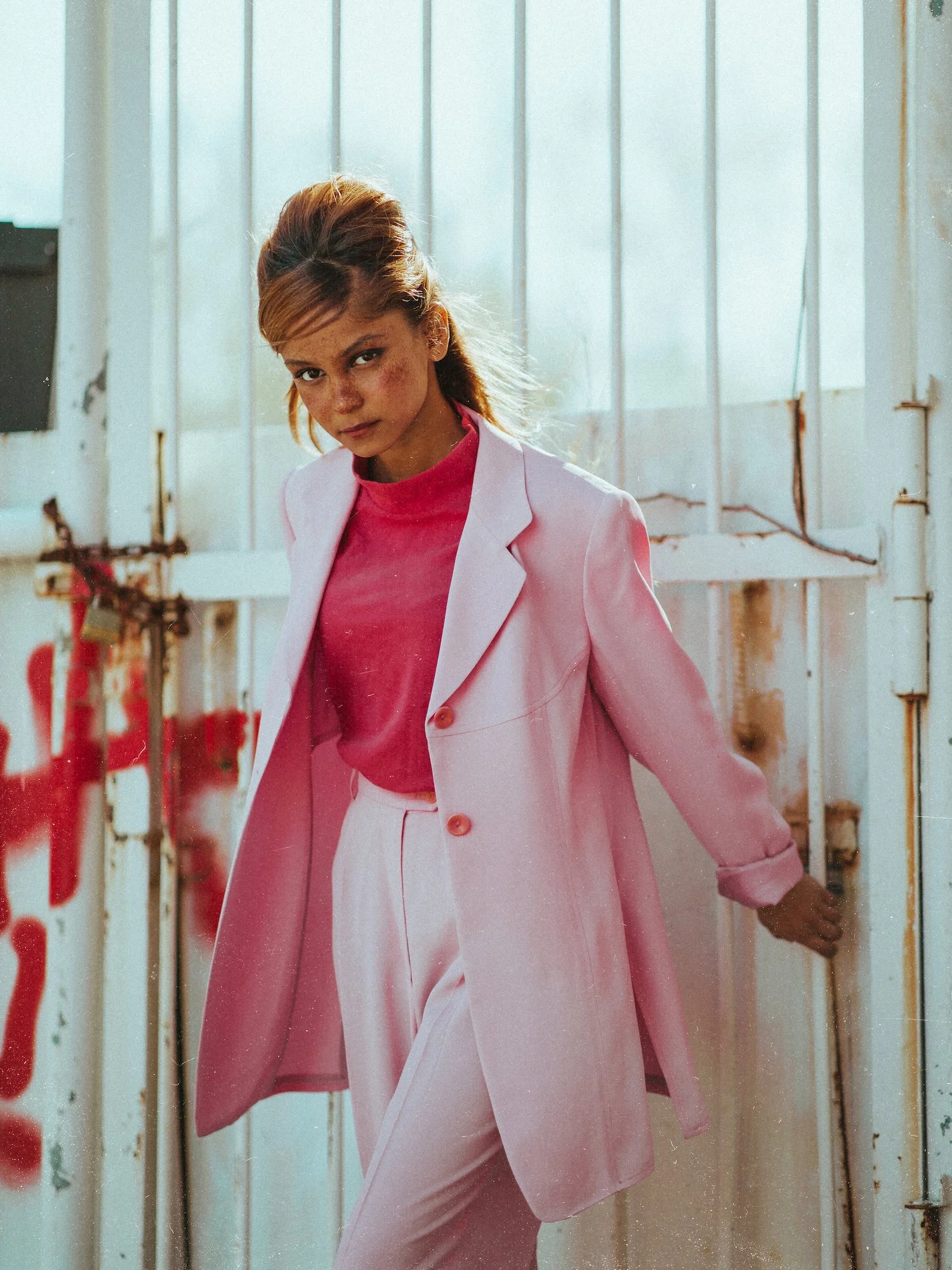 A woman in a pink suit and pink shirt standing in front of a rusty door with graffiti, looking at the camera.
