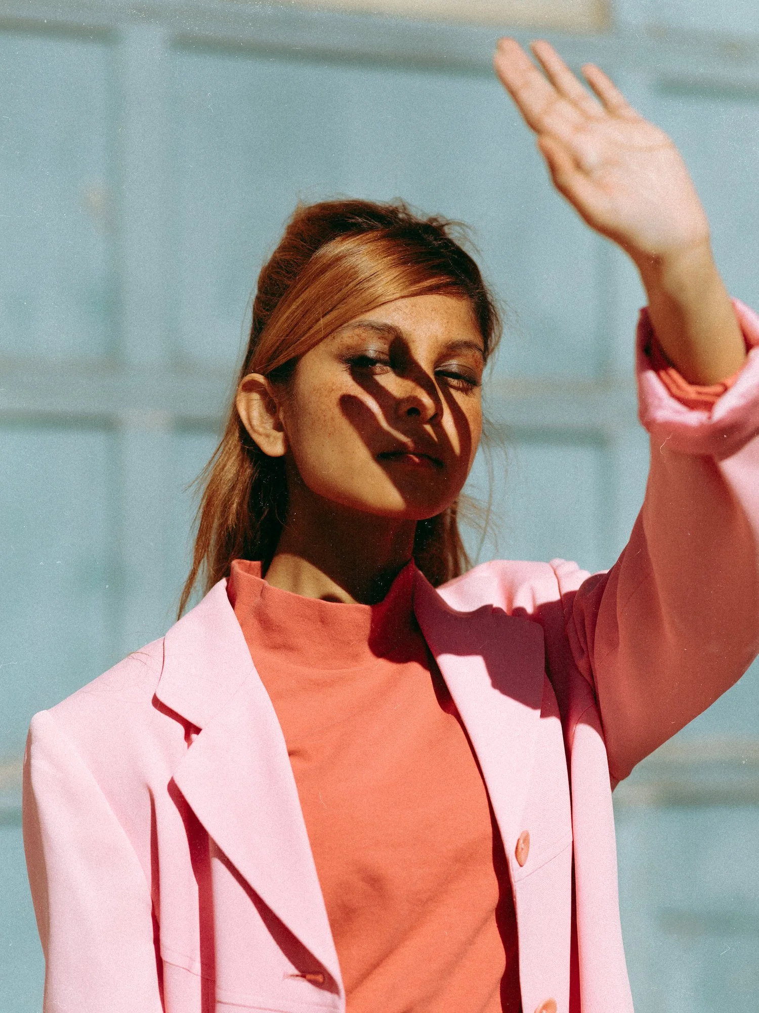 A woman with red hair is raising her hand, shielding her face from the sunlight, which creates a shadow of her hand on her face. She is wearing a pink blazer and an orange top, with a light blue background.