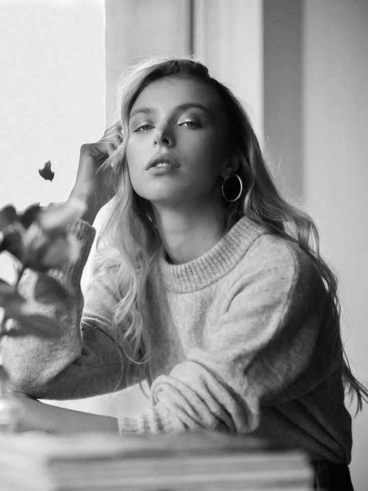 A black and white photo of a young woman with long wavy hair, wearing hoop earrings and a knitted sweater, sitting near a window with her hand resting on her head.