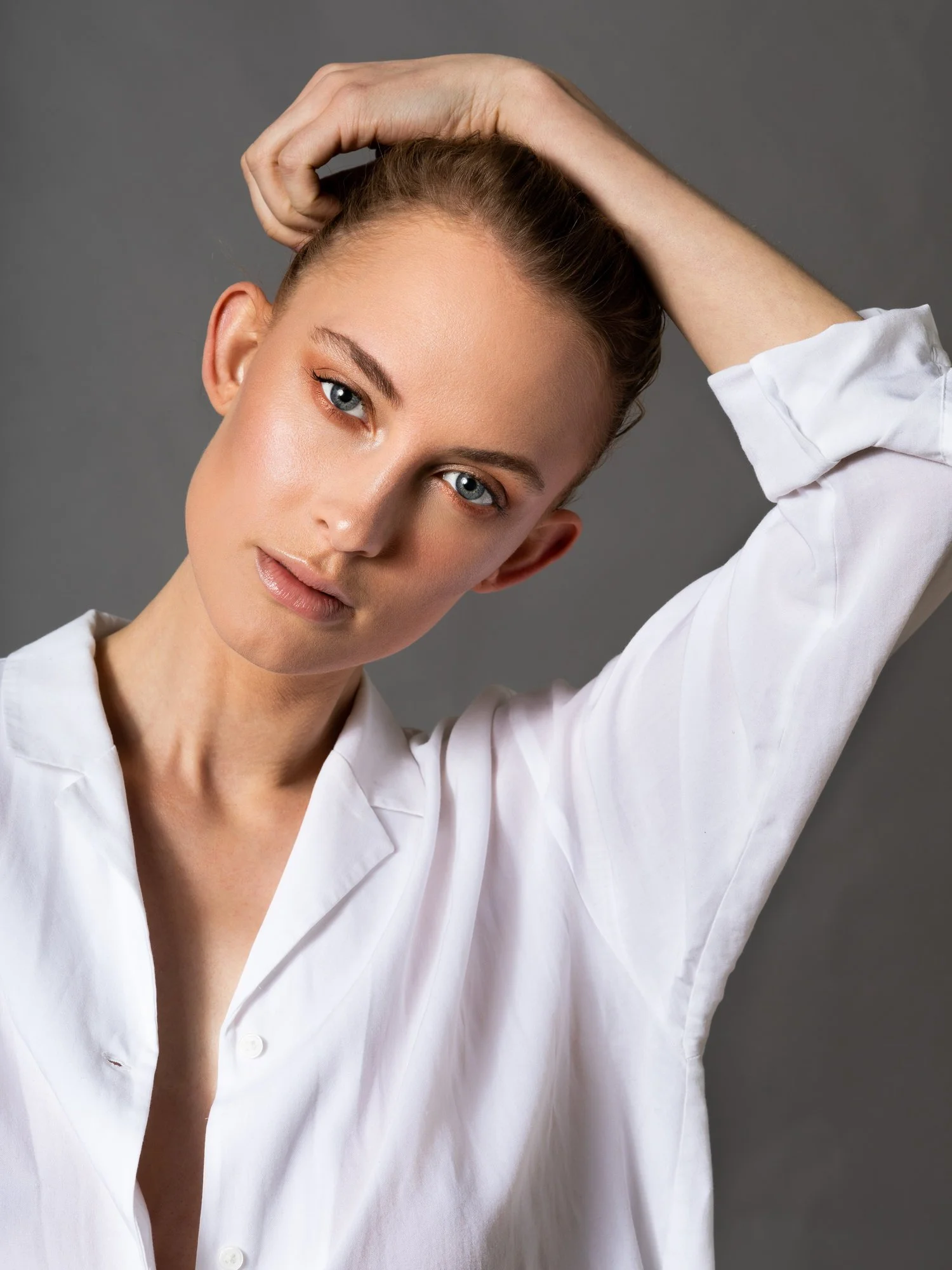 A young woman with blonde hair pulled back, wearing a white button-up shirt, is posing with one arm raised behind her head against a gray background.