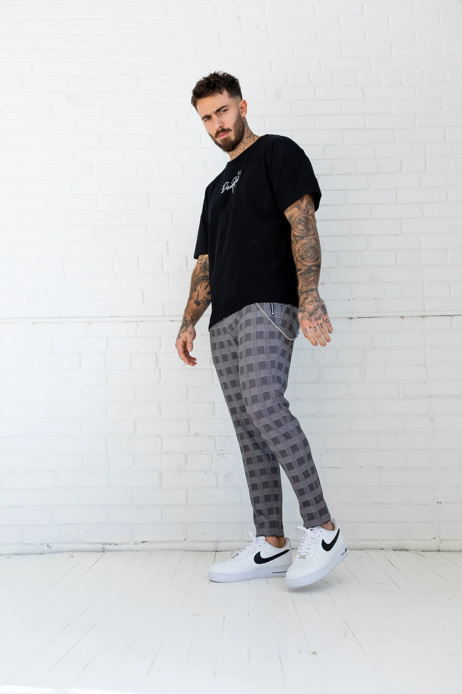 A tattooed man with a beard and styled hair wearing a black T-shirt, plaid pants, and white Nike sneakers, standing against a white brick wall.