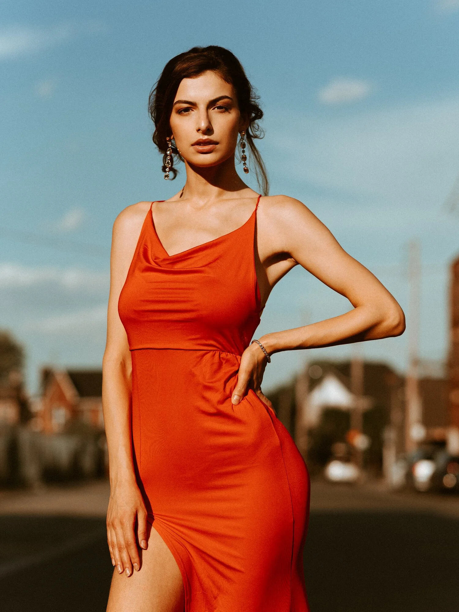 A woman in a red dress posing outdoors against a blue sky with some clouds, with blurred houses and cars in the background.