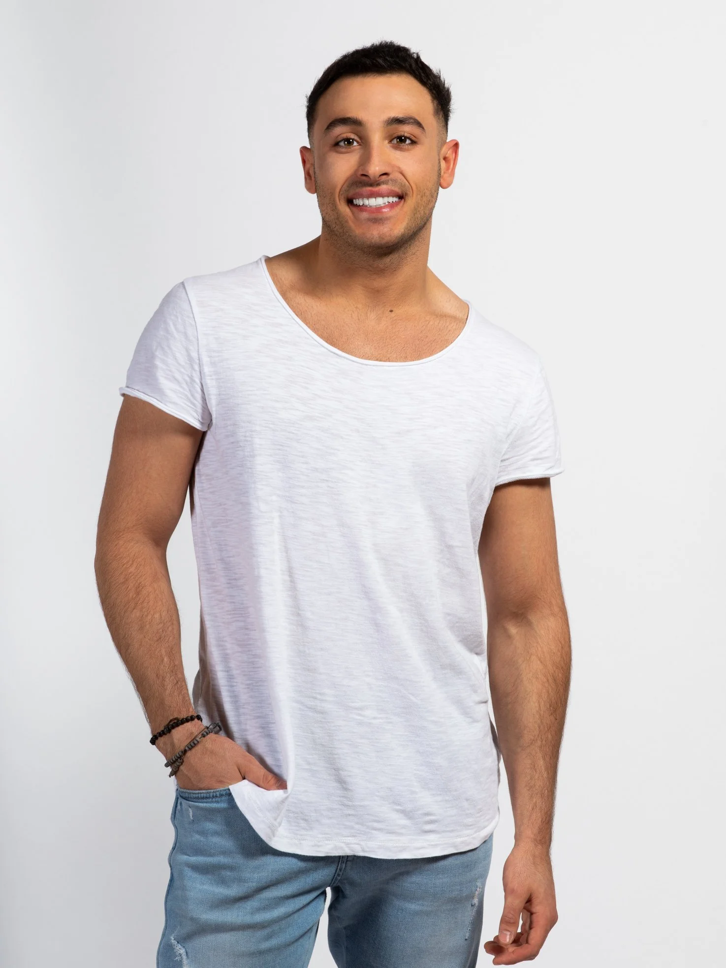 A young man with short dark hair and a light beard smiling, wearing a white t-shirt and light blue jeans, standing with one hand in his pocket against a plain white background.