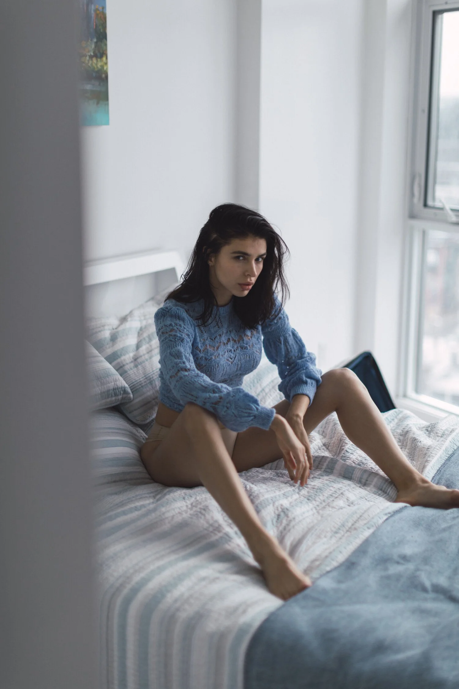 A woman with dark hair sitting on a bed by a window, wearing a blue knit sweater and beige shorts, with a serious expression.