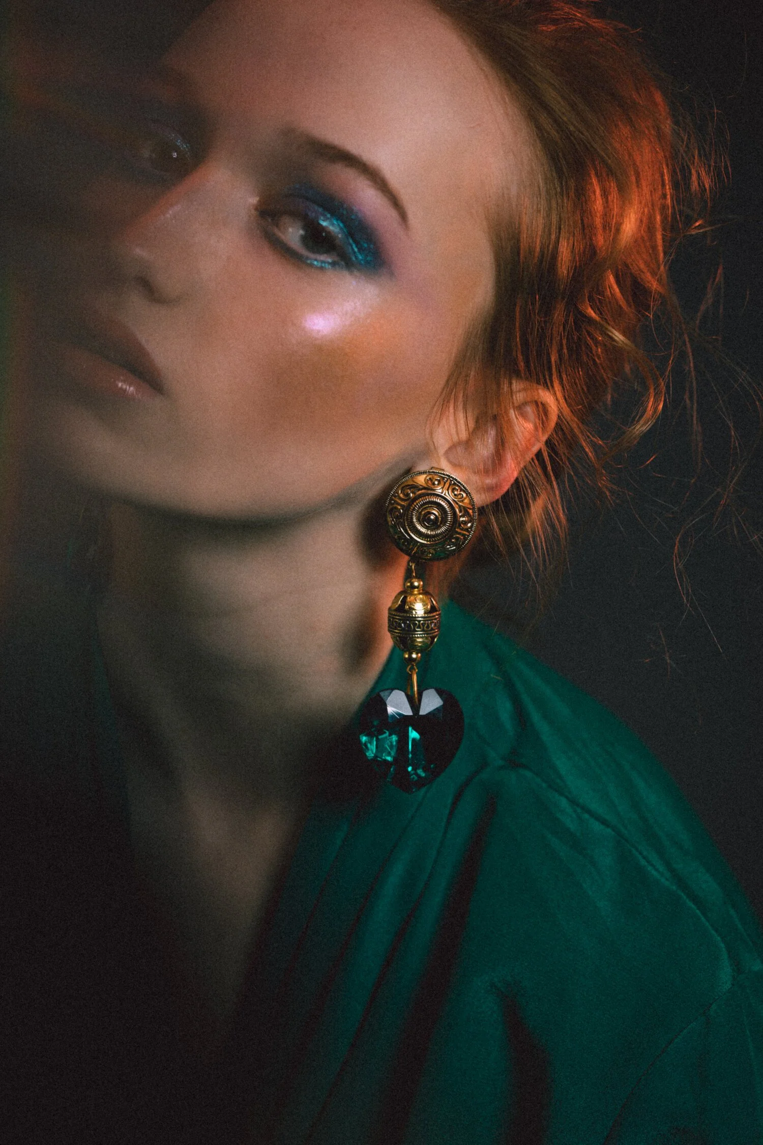 A close-up portrait of a woman with styled red hair, wearing bold blue eye makeup, shiny lip gloss, and a large ornate earring with a black heart-shaped pendant in a dark setting.
