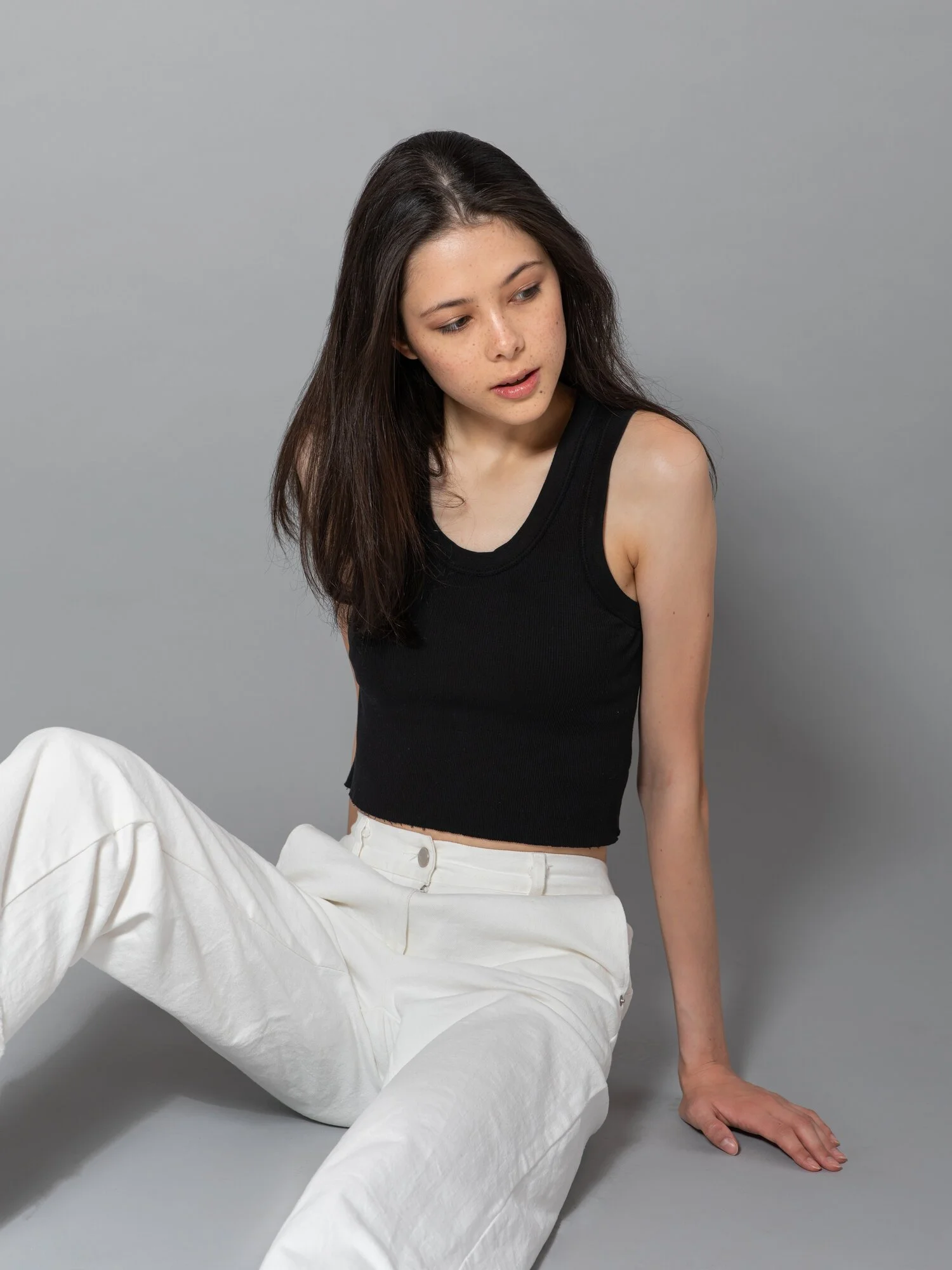 A young woman with long dark hair, dressed in a black sleeveless crop top and white pants, sitting on the floor against a plain gray background, looking to her left.