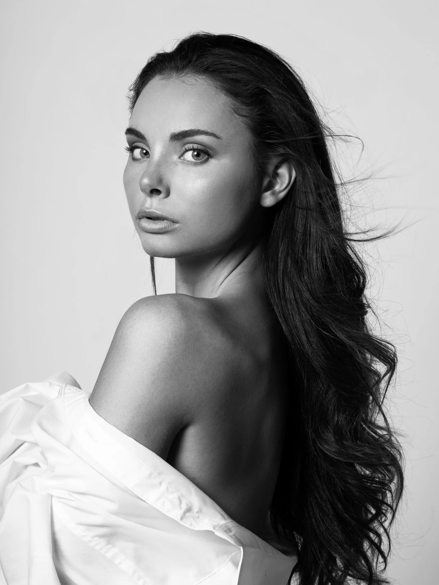 Black and white portrait of a woman with long, wavy hair, looking over her shoulder with a serious expression, partially covered by a white shirt.