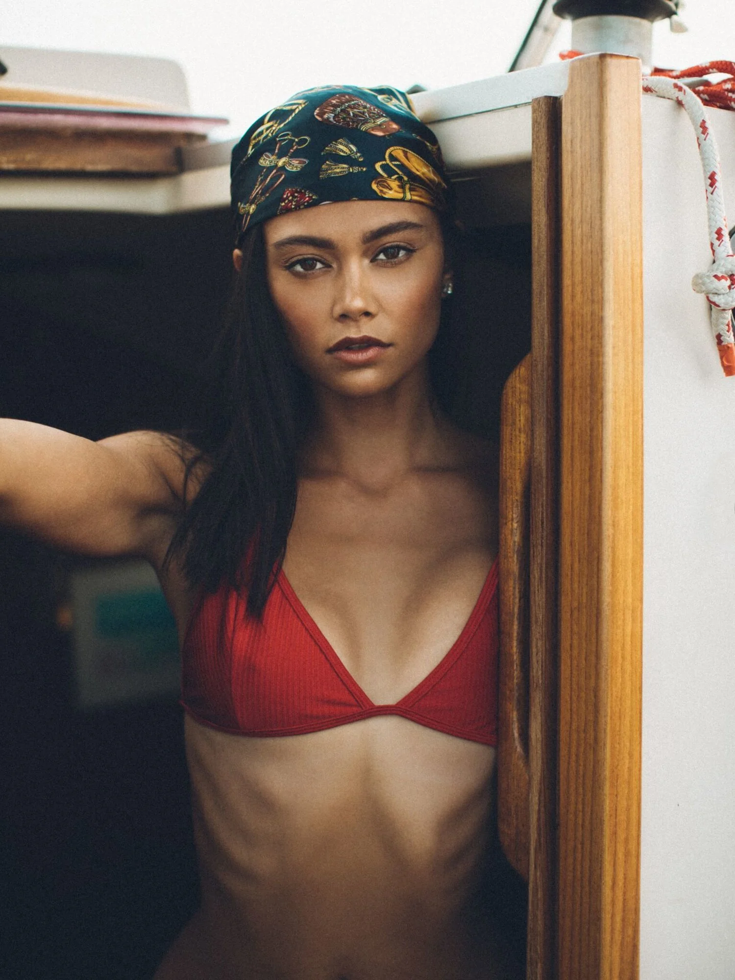 A woman in a red bikini top and a colorful headscarf poses under a table, looking directly at the camera with a serious expression.