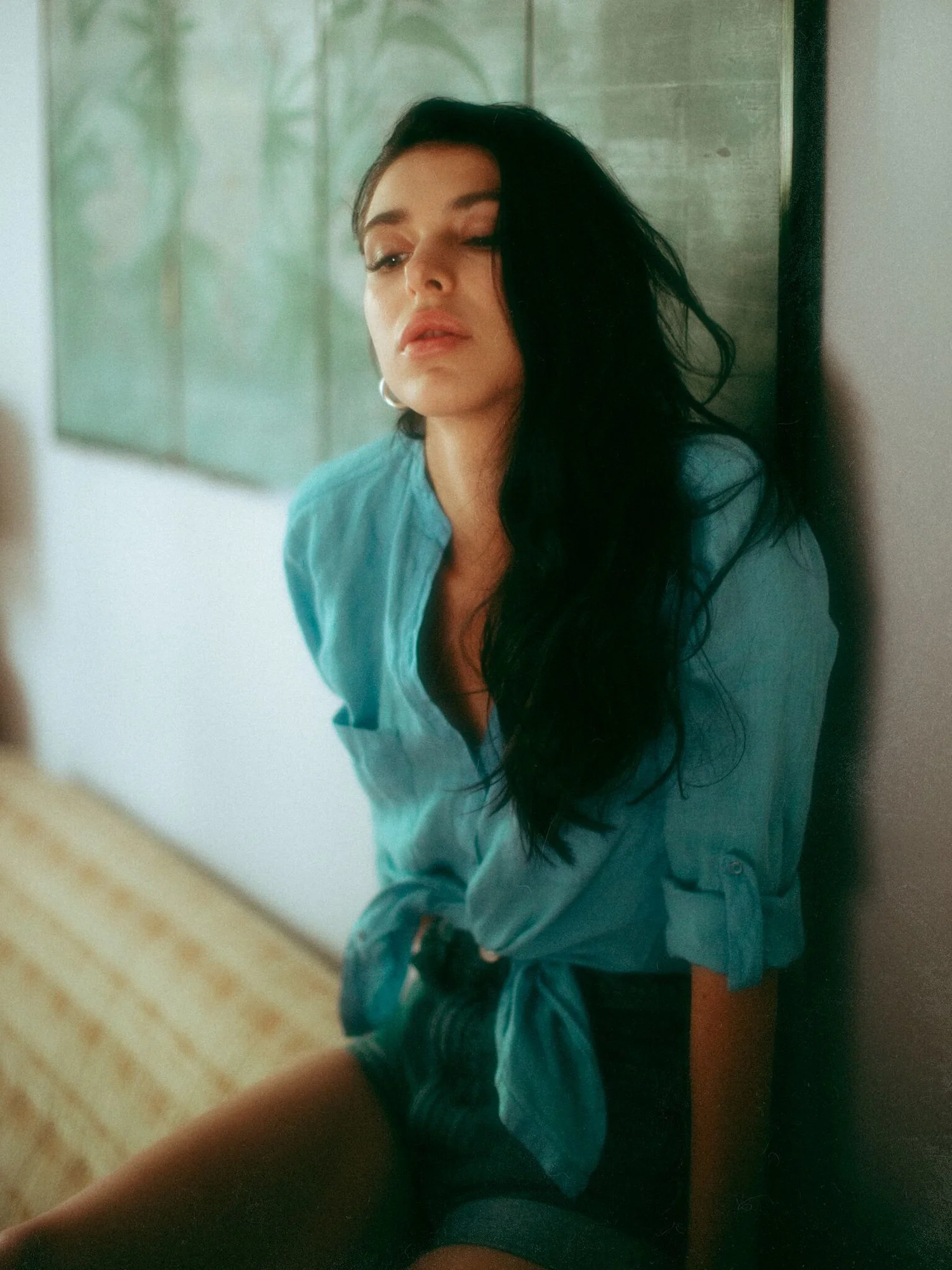 A woman with long black hair, wearing a blue shirt with rolled-up sleeves, sitting against a wall with a green landscape painting in the background.