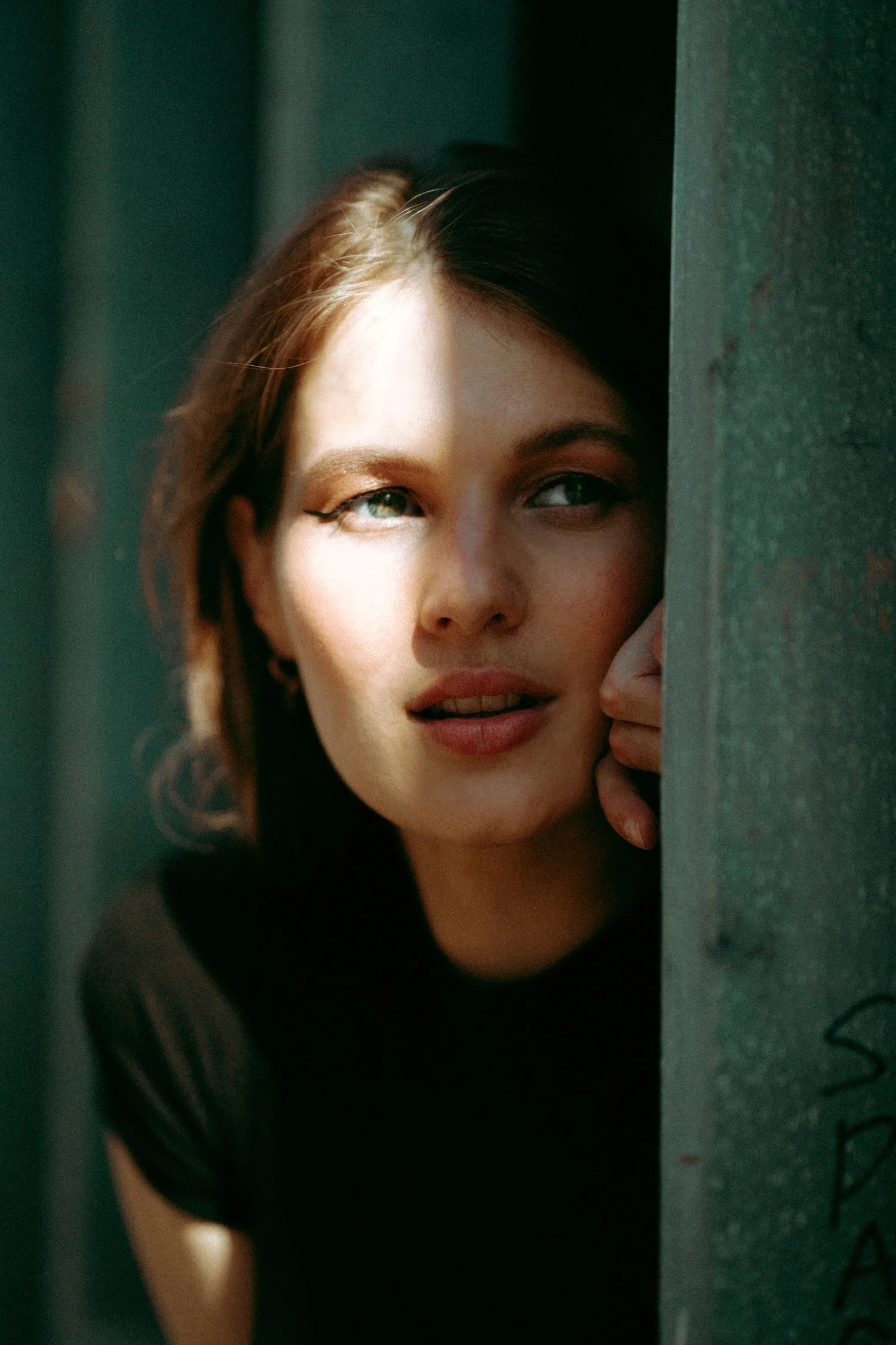 A woman with short hair and makeup peeks from behind a wall, sunlight casting a shadow across her face.