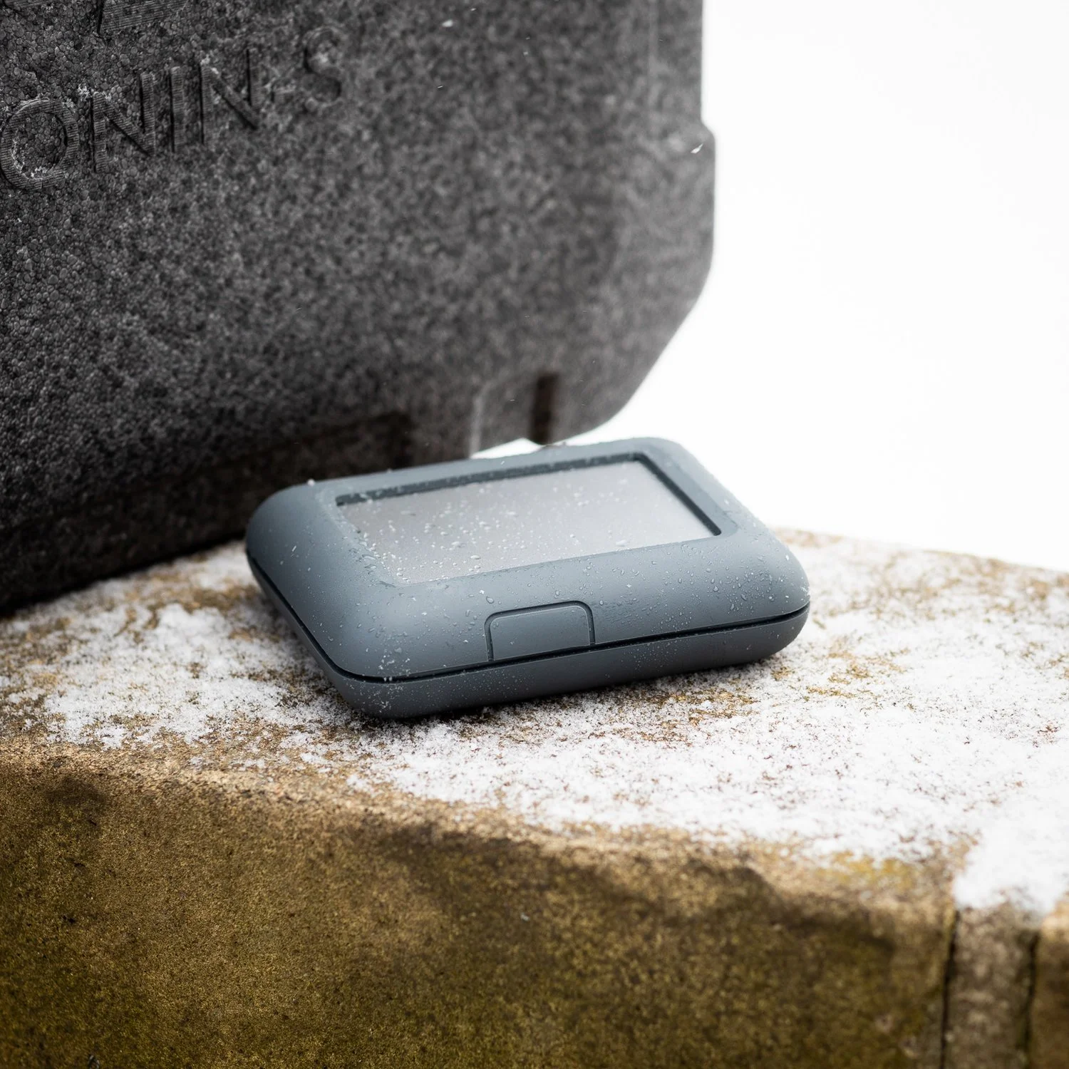 A gray GPS device with a rectangular screen resting on a snow-dusted brick surface outside, with a black textured surface in the background.
