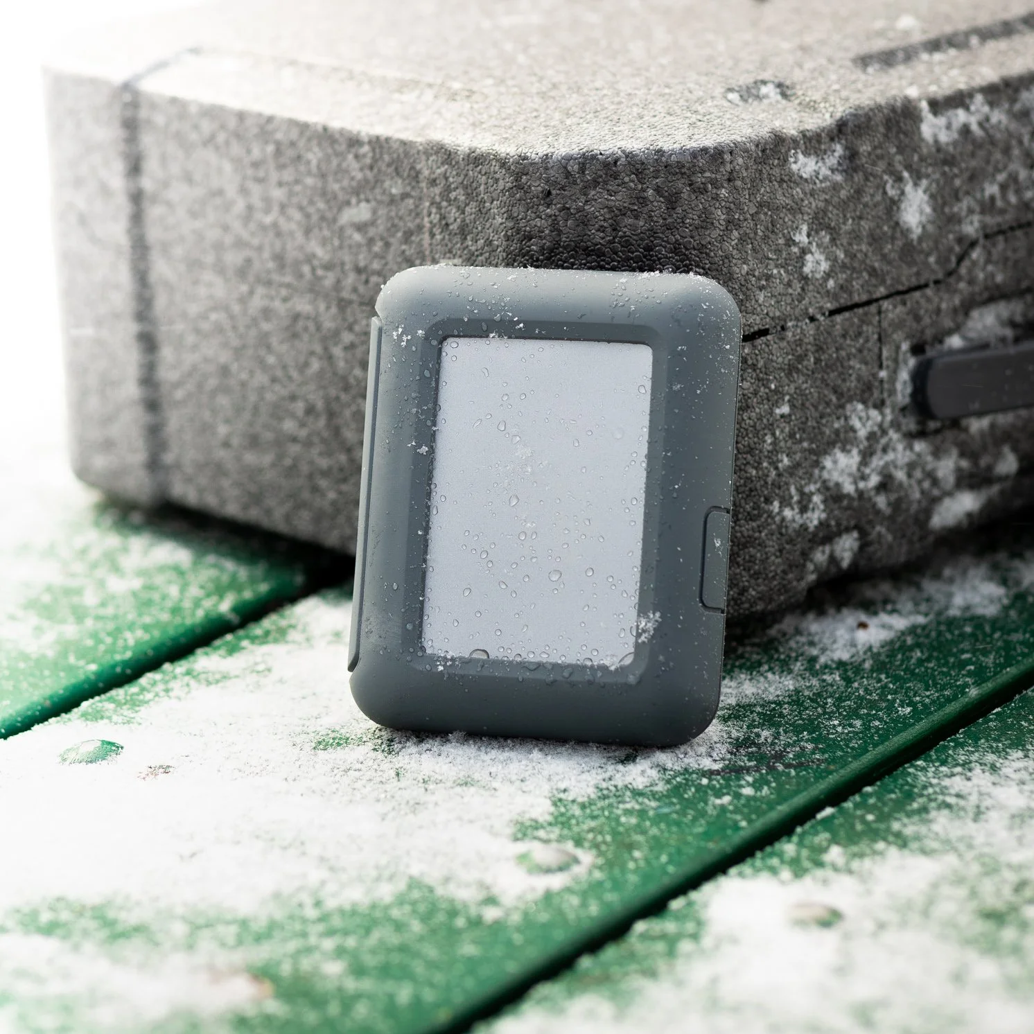 A digital watch with a gray case and water droplets on the screen, leaning against a gray, textured, snow-dusted surface outdoors.