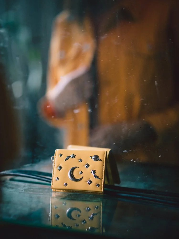 A yellow handbag with celestial-themed metal embellishments, including stars, moons, and planets, sitting on a reflective surface in front of a blurred background that appears to be a face in a mirror.