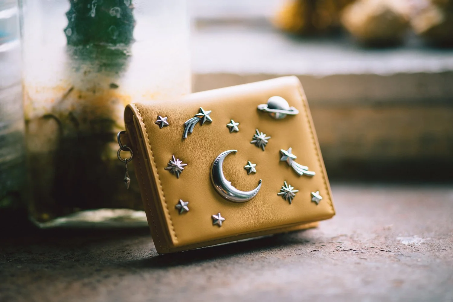 A tan wallet decorated with silver celestial-themed charms including stars, moons, and planets, resting on a wooden surface near a glass jar with plants inside. In the background, there is a blurred outdoor scene with water and trees.
