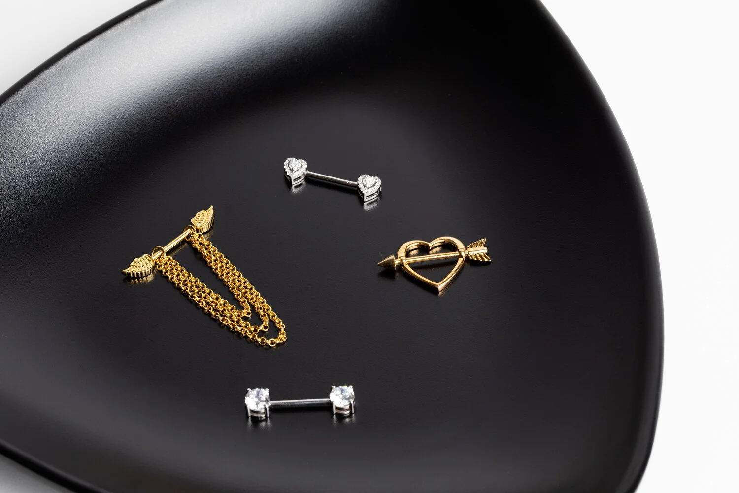 Set of four jewelry pins on a black surface, including a gold arrow with chains, a silver heart with an arrow, and two silver bar pins with diamonds.