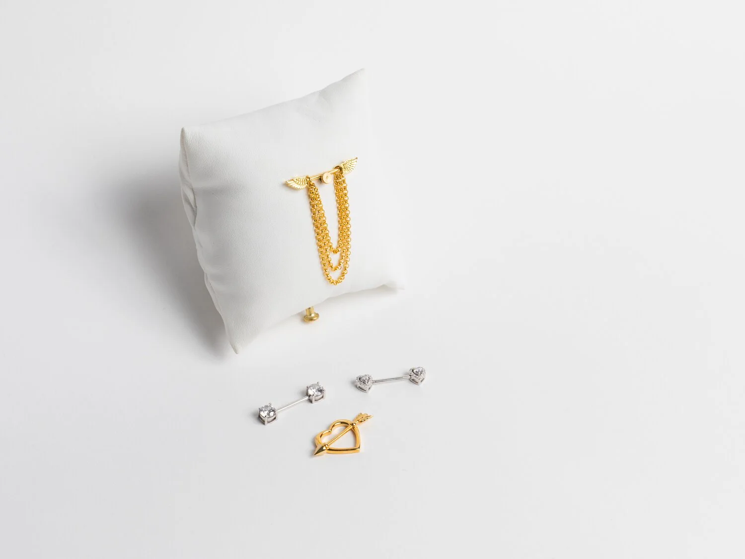Gold necklace with winged charm displayed on white pillow, with matching earrings and a heart-shaped pin with arrow in front.