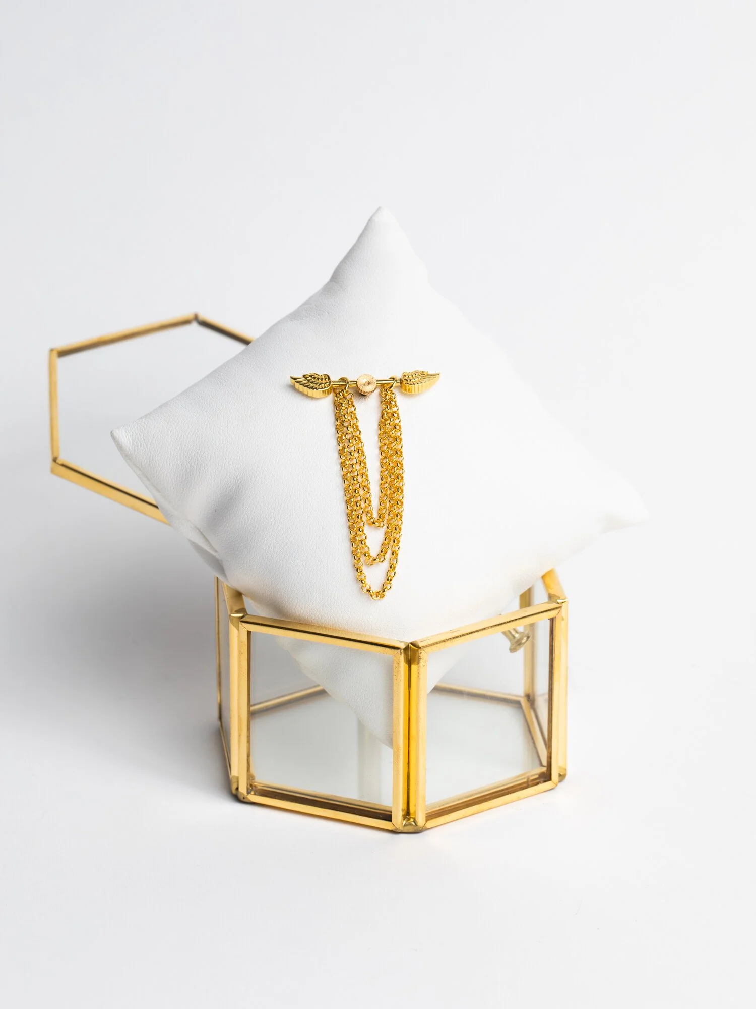 Gold jewelry piece with chains and wings on a white pillow inside a glass and gold box.