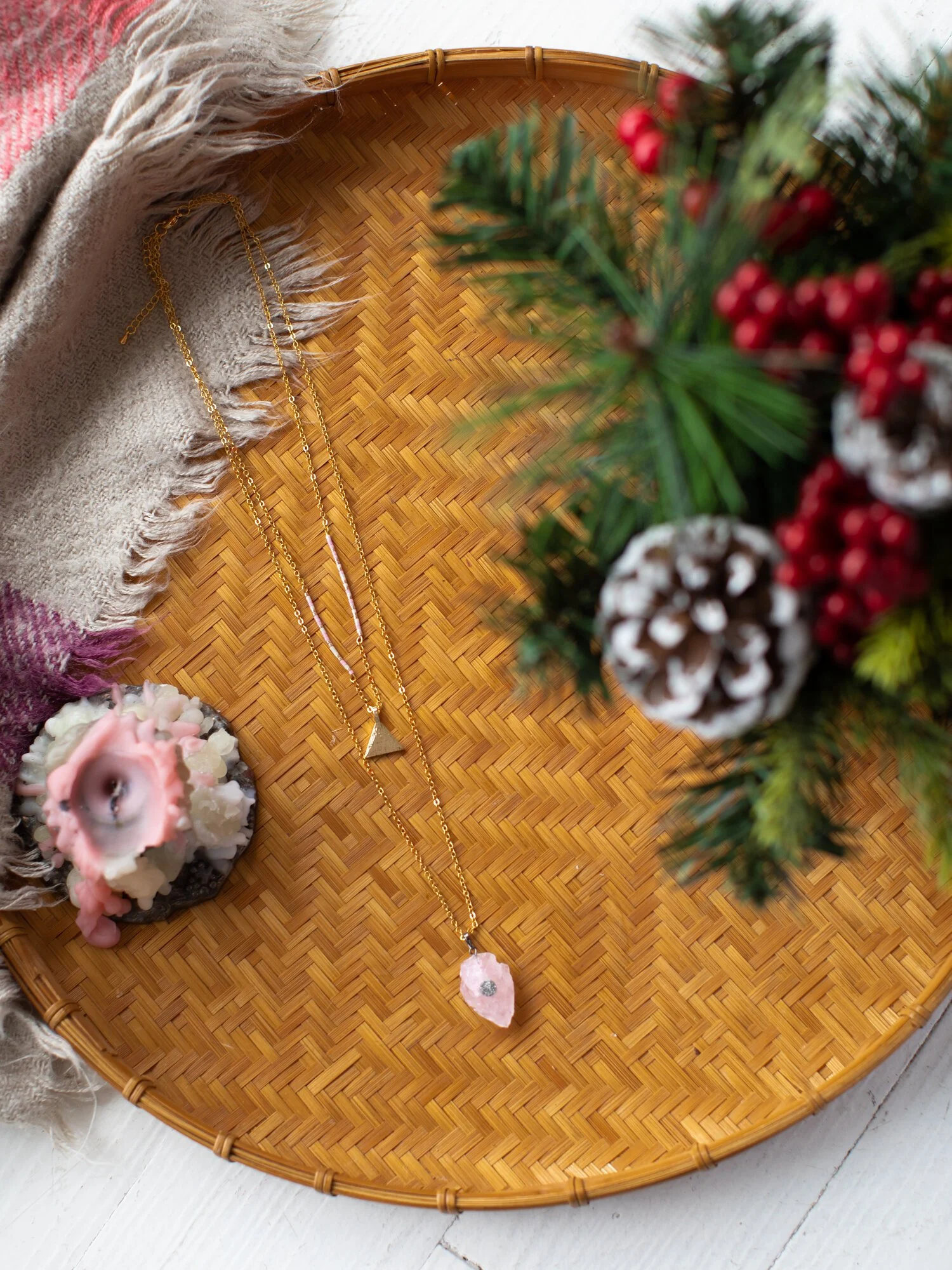 Golden necklaces with pink gemstone pendants arranged on a woven wooden tray, surrounded by a beige fringed scarf, pink and white candles, and a holiday-themed floral arrangement with pinecones and red berries.