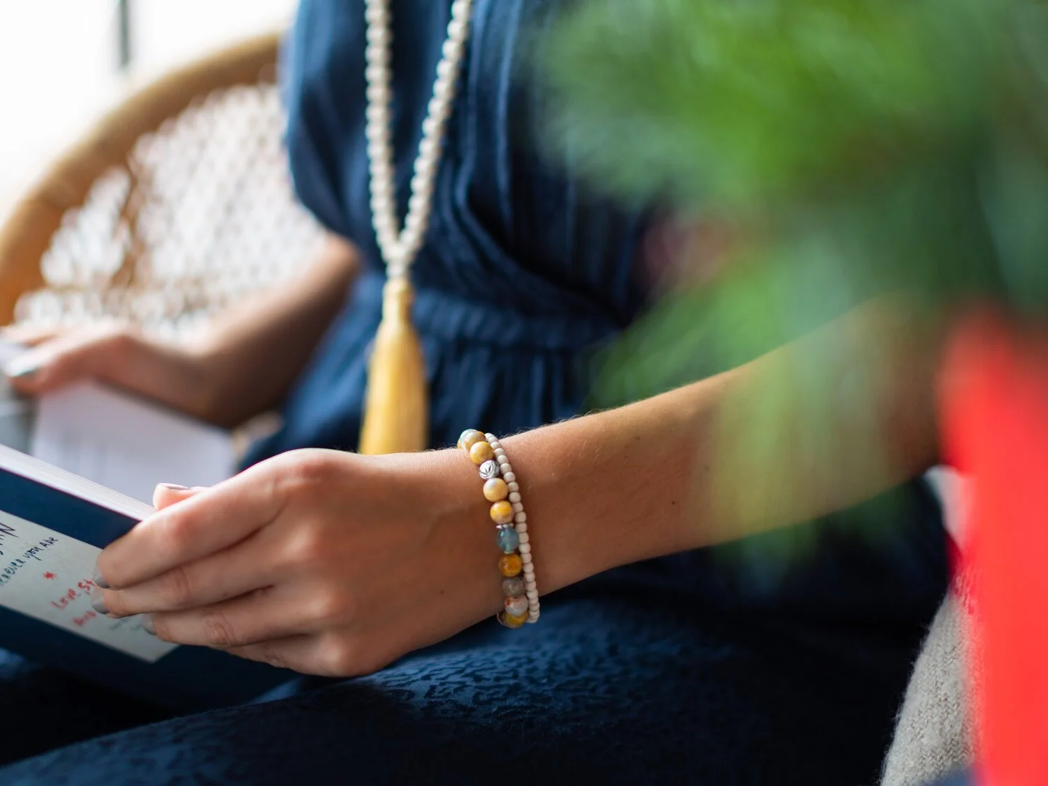 Close-up of a person's hand holding a book. The person is wearing a navy blue dress and accessorized with a string of white and yellow beads, a bracelet with multicolored beads, and a long beaded necklace with a yellow tassel.