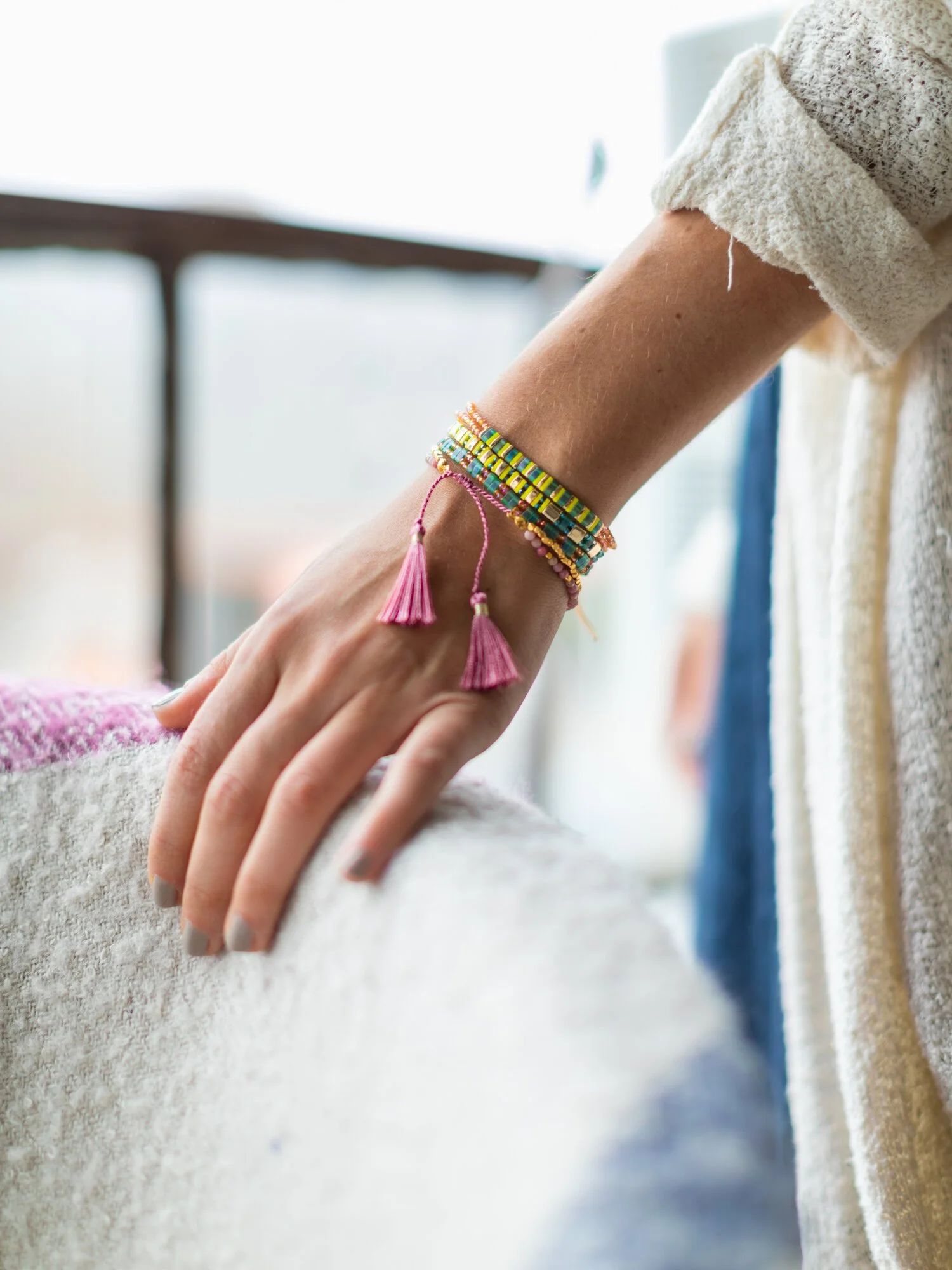 Close-up of a woman's hand with multiple colorful beaded bracelets and pink tassel bracelets resting on a textured white surface, with a blurred urban background seen through a window.