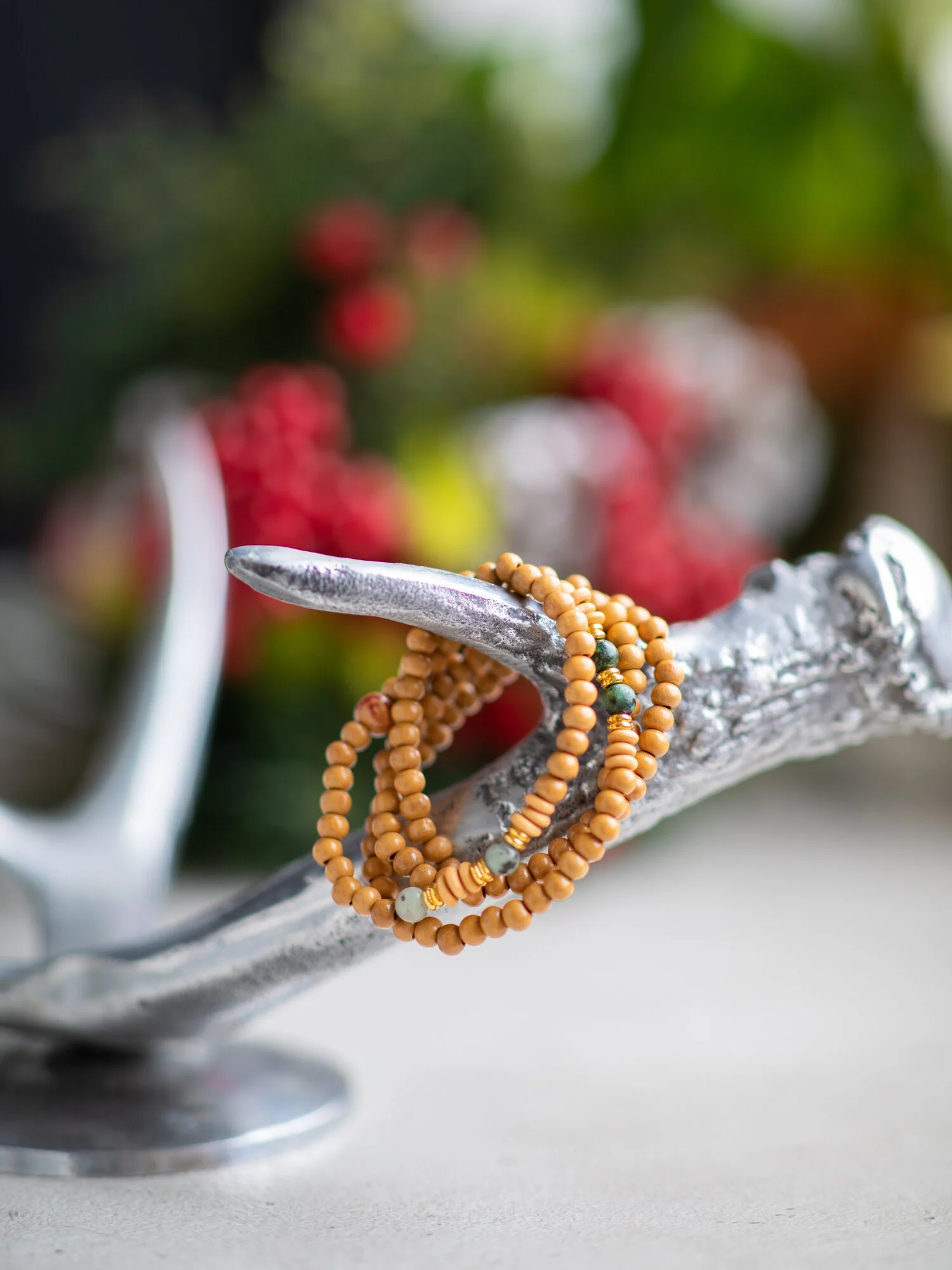 Beaded jewelry draped over a silver tree branch with red, white, and yellow flowers blurred in the background.