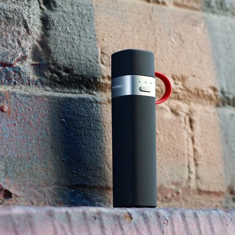 A portable black and gray coffee thermos with a red handle, leaning against a brick wall.