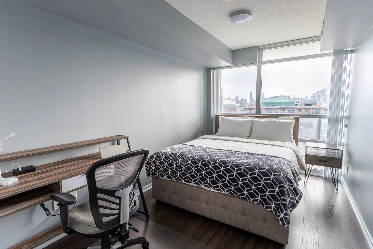 A modern bedroom with a bed topped with white linens and a black-and-white patterned blanket, a side table near a large window with city skyline view, and a wooden desk with a chair positioned against the wall.