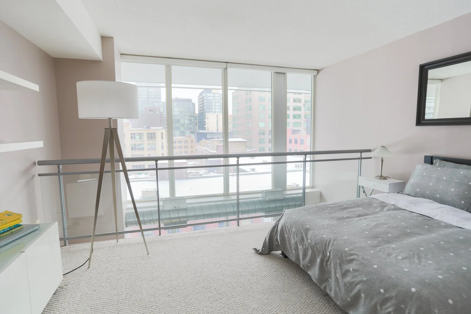 A modern bedroom with large floor-to-ceiling windows overlooking a city skyline, a bed with gray polka dot bedding, a white side table with a lamp, and a large mirror on the wall.
