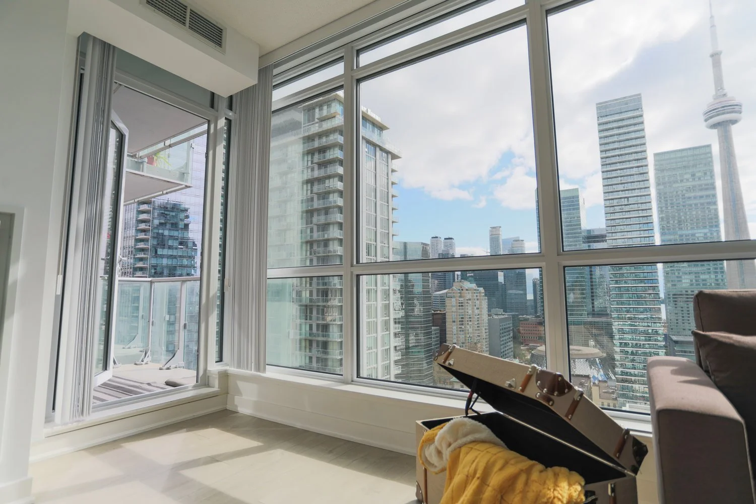 A modern city apartment with large floor-to-ceiling windows overlooking a city skyline with tall skyscrapers. Partially open balcony door and a chest with a blanket inside are visible.