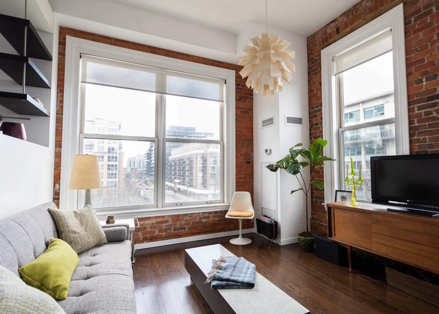 Modern living room with large windows, exposed brick walls, a beige sofa with colorful pillows, a wooden coffee table with a plaid blanket, a white chair, a tall potted plant, a TV on a wooden stand, a yellow candle holder, and a unique white hanging light.